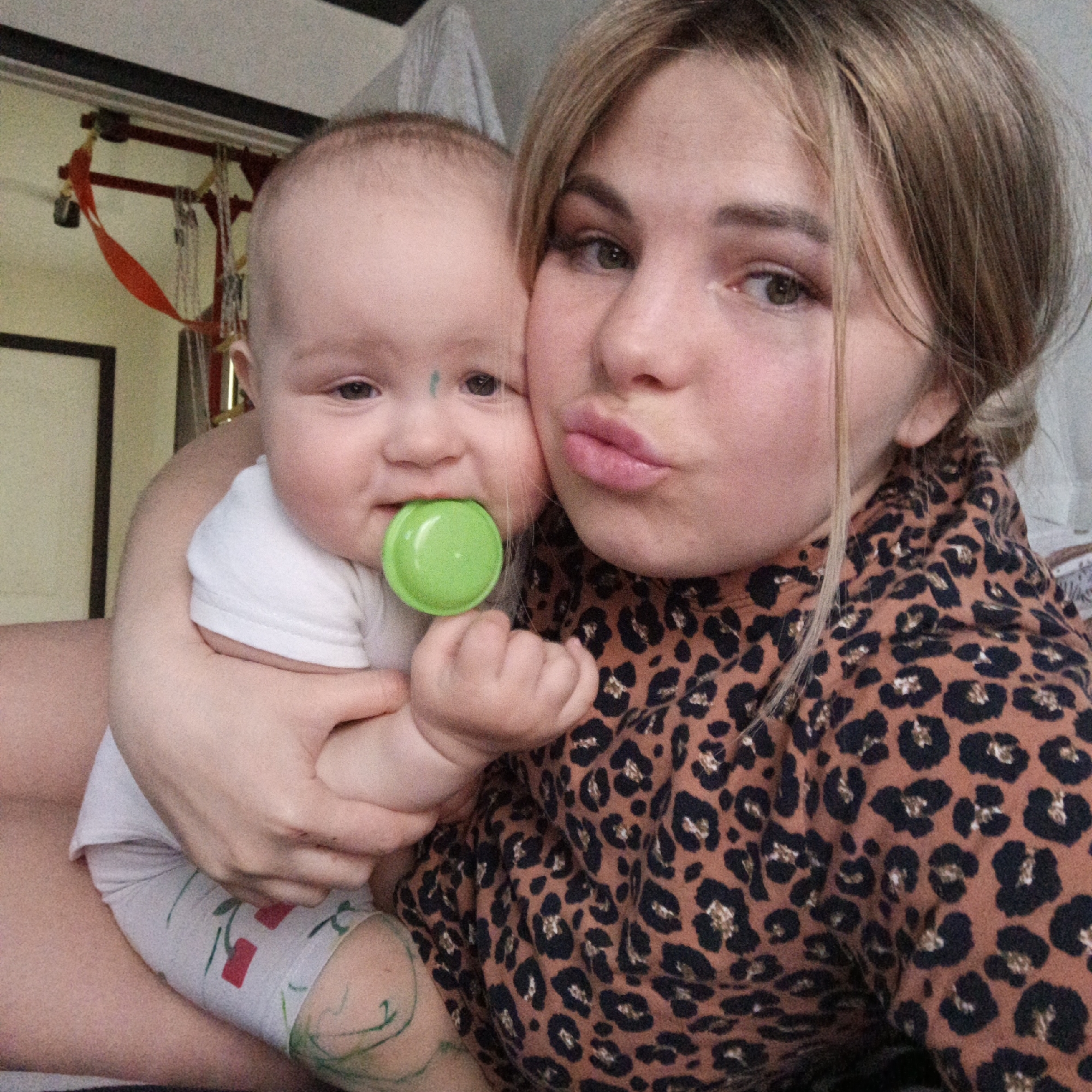 A young woman is holding a baby with a green pacifier. The woman is wearing a leopard print top and has blonde hair. The baby is wearing a white shirt and grey pants with cherries. The background shows a wall and some objects on a shelf. The location appears to be indoors.