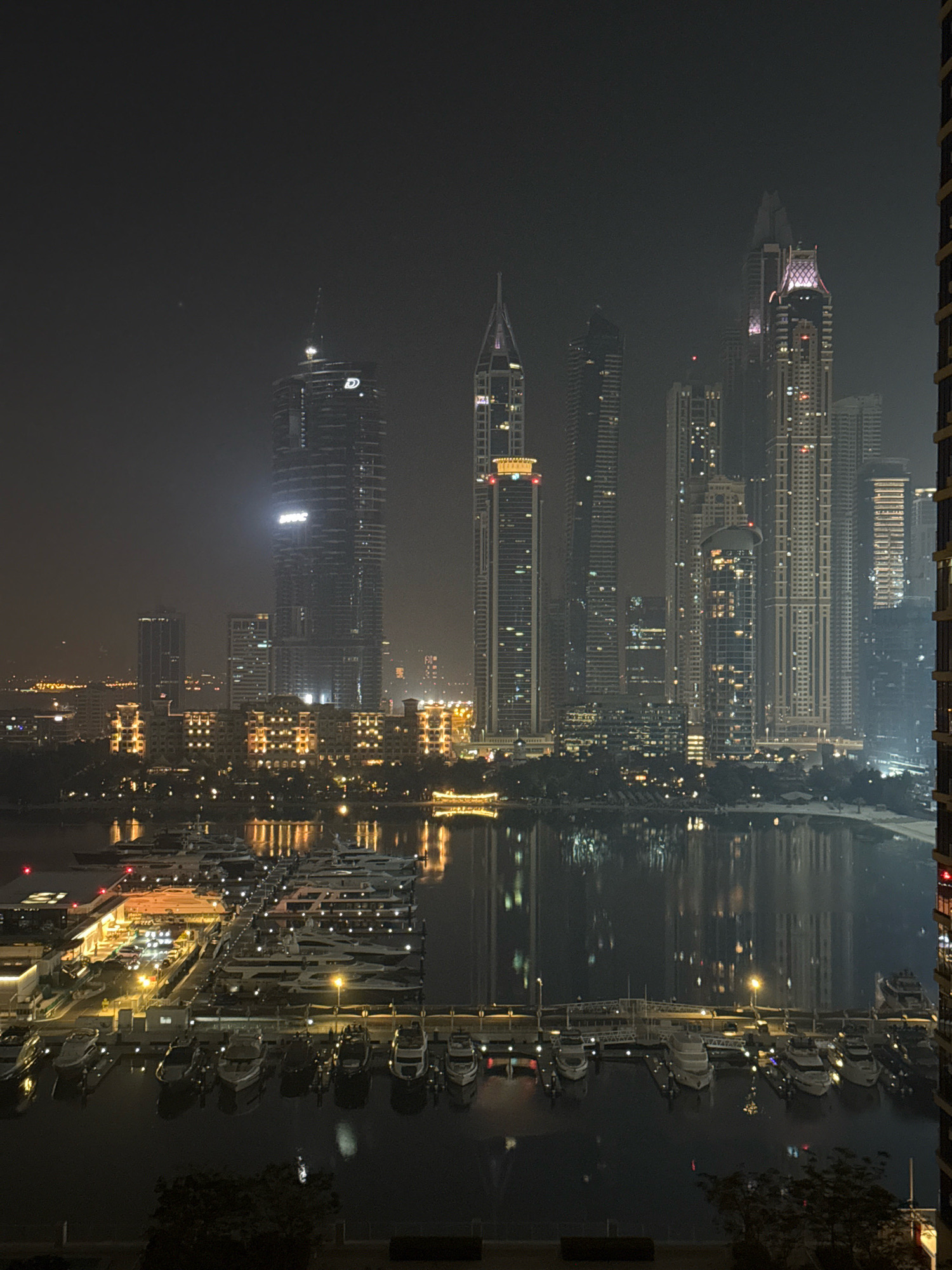 The image captures a nighttime panorama of a modern cityscape with a focus on a marina filled with yachts. Tall skyscrapers, illuminated by numerous lights, dominate the background, reflecting in the calm waters of the marina. The architectural style of the buildings suggests a contemporary urban environment, typical of Dubai.
In the foreground, numerous yachts of varying sizes are docked in neat rows. The marina is well-lit with artificial lights, highlighting the sleek designs of the vessels and the infrastructure surrounding them. The water's surface shows reflections of the city lights and the yachts, adding to the visual depth of the scene.
The sky is dark and appears slightly hazy, common in urban environments at night, and the overall lighting suggests it is late evening or night. The presence of advanced architecture and a sophisticated marina indicates a location known for luxury and modern development. Considering these elements, the scene is highly indicative of Dubai Marina, Dubai, United Arab Emirates. No people are discernibly visible in the image.