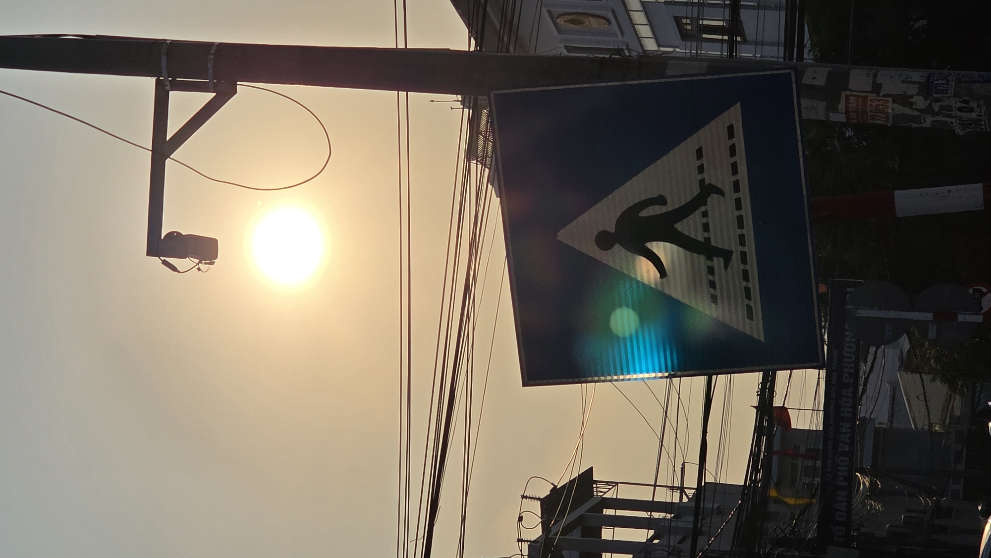 Here's a forensic description of the image:
The image shows a street scene in Bắc Nha Trang, Vietnam, taken during the late afternoon or early morning, as evidenced by the bright, low sun in the background. The perspective is from below, looking upwards.
A pedestrian crossing sign is prominently displayed, featuring a blue background with a white triangle containing a black silhouette of a walking figure. The sign is mounted on a pole amidst a dense network of overhead wires. A camera is attached to a pole near the sun.
Behind the crossing sign, a building is partially visible, along with a pole plastered with posters, likely advertisements or notices. A red and white barricade is also visible in the background. A sign is also visible, which has the text "TỔ DÂN PHỐ VĂN HÓA PHƯƠNG, 2053", indicating an address or district of the city.
The overall environment appears to be urban and densely populated, with a mix of infrastructure and signs. The sky is washed out due to the bright sunlight.