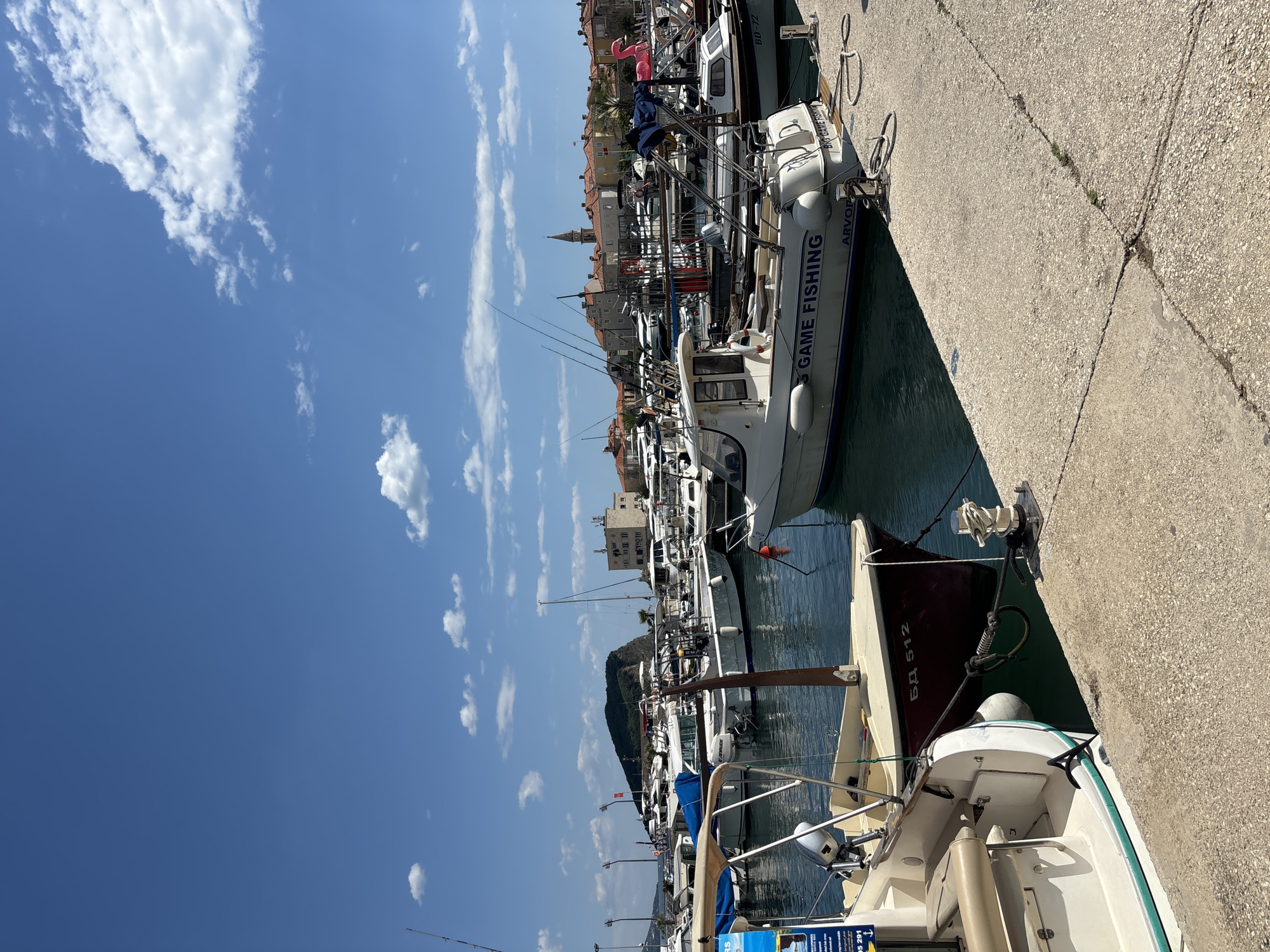 The image captures a bustling marina in Budva, Montenegro, with a clear blue sky dotted with fluffy white clouds overhead. Numerous boats of varying sizes and types are moored along the concrete quay. A white motorboat with the words "GAME FISHING" on its side is prominently featured. Other details visible include fishing rods extending upwards, the tops of buildings with red-tiled roofs in the background, and a partially obscured boat with "БД 512" and "05 291" painted on its stern. The concrete quay shows signs of weathering and cracks. The water in the marina appears calm. The scene suggests a vibrant harbor with activity related to boating and fishing.