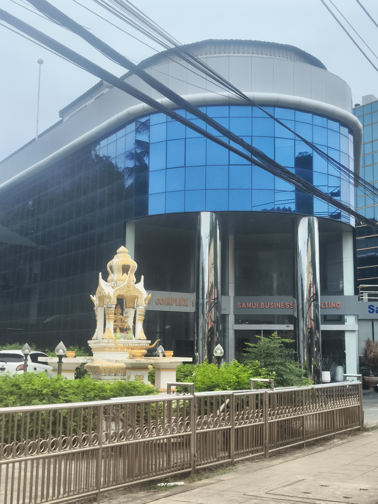 This image shows a modern building with a curved facade of blue glass windows, identified as "SAMUI BUSINESS" and "COMPLEX" by signage. In front of the building is a decorative, ornate shrine with golden accents, flanked by lush green bushes. A metallic fence runs along the foreground, and power lines are visible crisscrossing the sky. To the left, a white car is parked near the entrance of the building. The overall scene is one of a commercial area in Baan Chaweng Noi, Thailand, characterized by contemporary architecture and cultural elements.