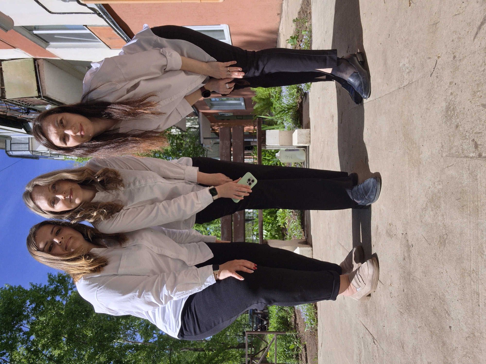 Three women are vertically aligned in an outdoor setting in an urban area. The top woman has long brown hair, a light-colored long-sleeved shirt, and black pants. She is grasping the waist of the woman below. The middle woman has blonde hair, a light-colored long-sleeved shirt, black pants, and is holding a smartphone with a green case. The bottom woman has brown hair, a light-colored long-sleeved shirt, and black pants, and is grasping the waist of the woman above. The background includes buildings with windows and a peach-colored wall, foliage, and a concrete surface.