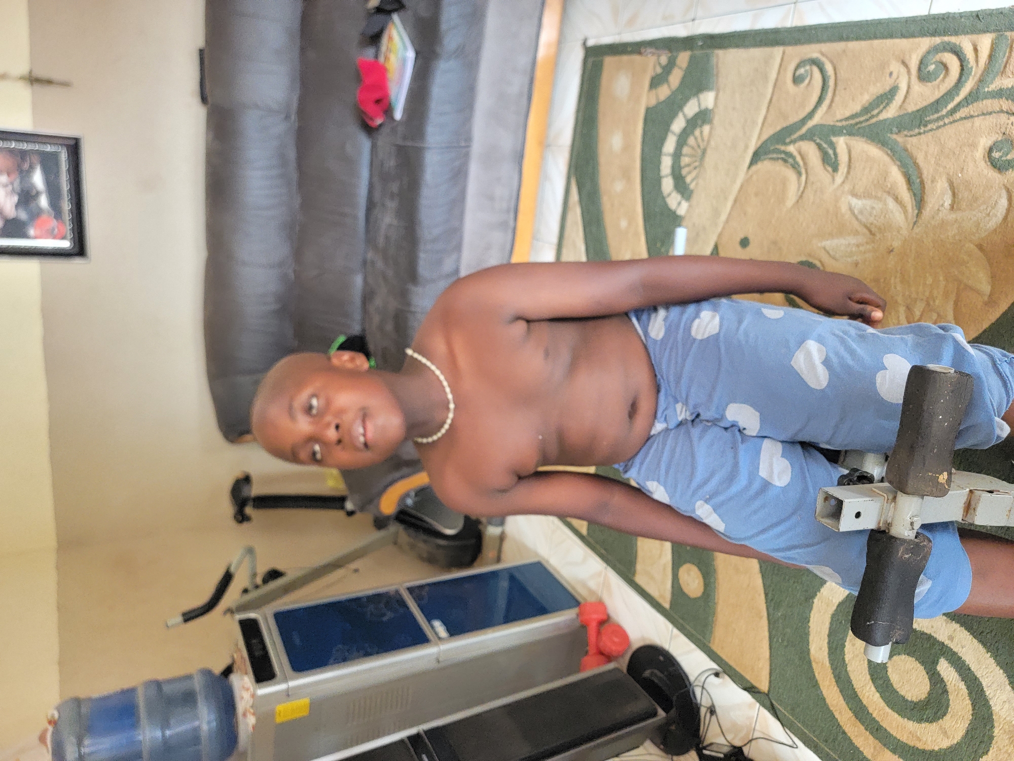 A young, dark-skinned, shirtless boy wearing light blue pajamas with white hearts and a pearl necklace is reclining on a leg extension weight machine on a patterned rug. The environment appears to be indoors, near a water cooler, a silver machine with blue water, and other workout equipment. A picture frame is hung on the pale yellow wall. There's no explicit evidence of the city, Sabon Tasha, Nigeria.