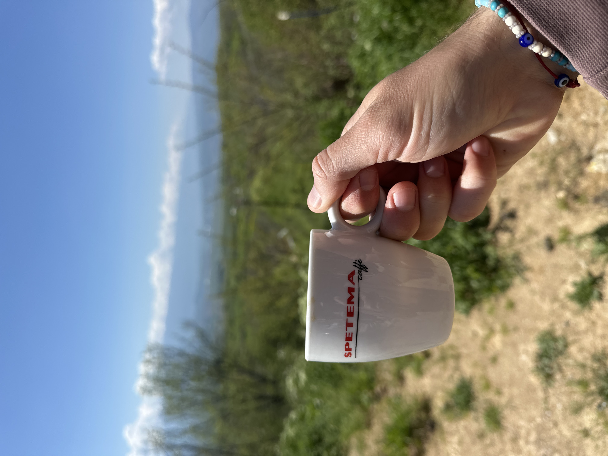 A person's hand holding a white coffee mug with "SPETEMA caffe" printed on it, with a landscape background. The foreground is dominated by the hand holding the mug. The wrist has two bracelets, one with turquoise, white, and blue beads, and another with a red cord and small blue beads.
The background appears to be a hilly landscape with vegetation, possibly trees and shrubs. In the distance, there's a hazy blue sky, suggesting a clear day. The angle of the photograph makes it difficult to determine the exact location. Based on the location you provided, the landscape aligns with the hilly terrain found in Bulgaria near Lyubovka. The image appears to have been taken outdoors in natural lighting.