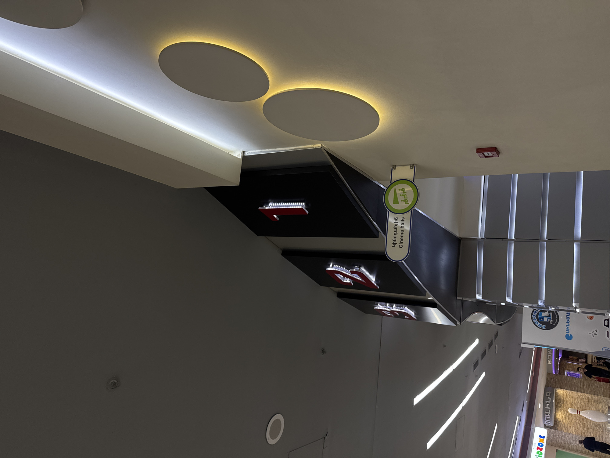 The image depicts the interior of a commercial building, potentially a shopping mall or entertainment complex, in Yerevan, Armenia. The scene includes:
- A sign indicating "Cinema Halls" in Armenian.
- Directional signage with arrows and illuminated logos of what might be cinema halls or theaters.
- Escalators or stairs leading to the upper level, illuminated with linear lights.
- Shop signs, including a "BOW" sign, visible in the distance suggesting the presence of bowling alleys.
- Individuals walking on the mall floor.
- Circular lights installed on the ceiling.
- Sprinkler system on the ceiling.
- Audio system component in the wall.