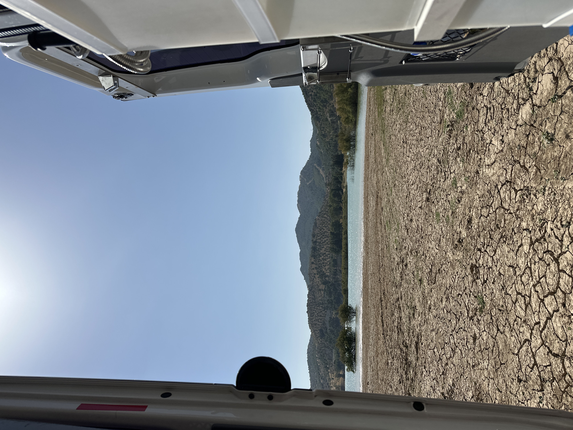 The photograph is taken from inside a vehicle, looking out at a dry, cracked earth landscape leading to a body of water. The foreground is dominated by the parched ground, with visible fissures and sparse, dry vegetation. To the left, the interior of a white vehicle is visible, including a black circular object and a red reflective strip on a white panel. To the right, the exterior of a gray and white vehicle is seen, with a metallic hose attachment and part of a white container.
In the midground, a large expanse of water reflects the bright sunlight, shimmering under a clear blue sky. The water's edge meets the dry land, and two small trees are partially submerged in it. Beyond the water, a series of rolling hills rise, covered in vegetation, with some appearing to be olive groves. The sky is a vibrant, cloudless blue, with the sun positioned high and slightly to the left, casting a bright light over the scene. The overall impression is one of a dry, arid environment adjacent to a lake, likely in Ardales, Spain, given the terrain and vegetation. No people are visible in the image.