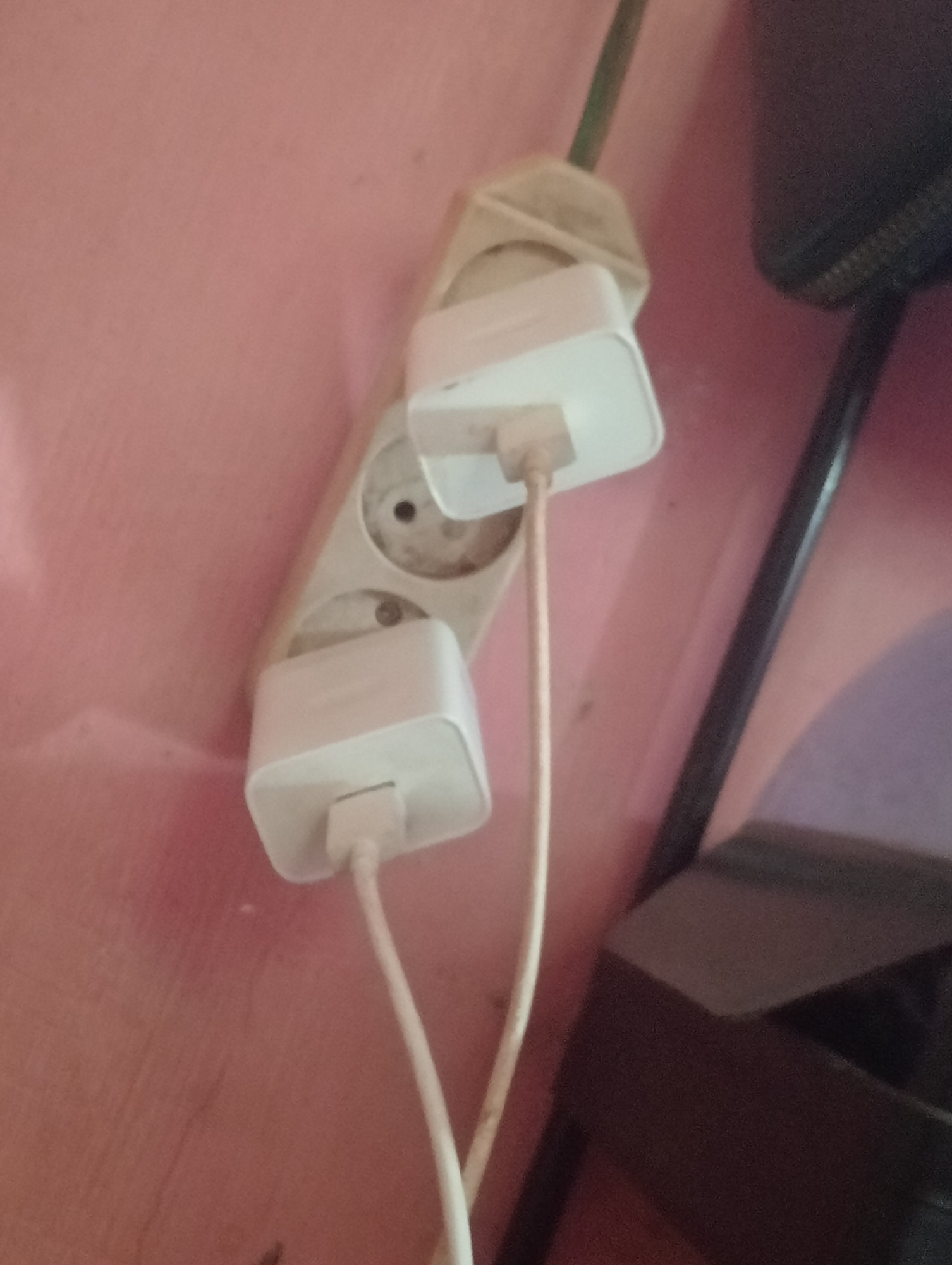The image shows a wall with a pink textured surface, likely painted. A white electrical extension cord with multiple outlets is plugged into the wall. Two white rectangular power adapters, possibly for charging electronic devices, are plugged into two of the outlets on the extension cord. White charging cables extend from these adapters. To the right of the extension cord, a dark object, possibly furniture or an electronic device with a textured surface, is visible. The background shows a slight gradient of color, from pink to a muted purple. The scene is indoors, with no indication of a specific location within Bloro, Indonesia, other than it is an indoor domestic or office setting.