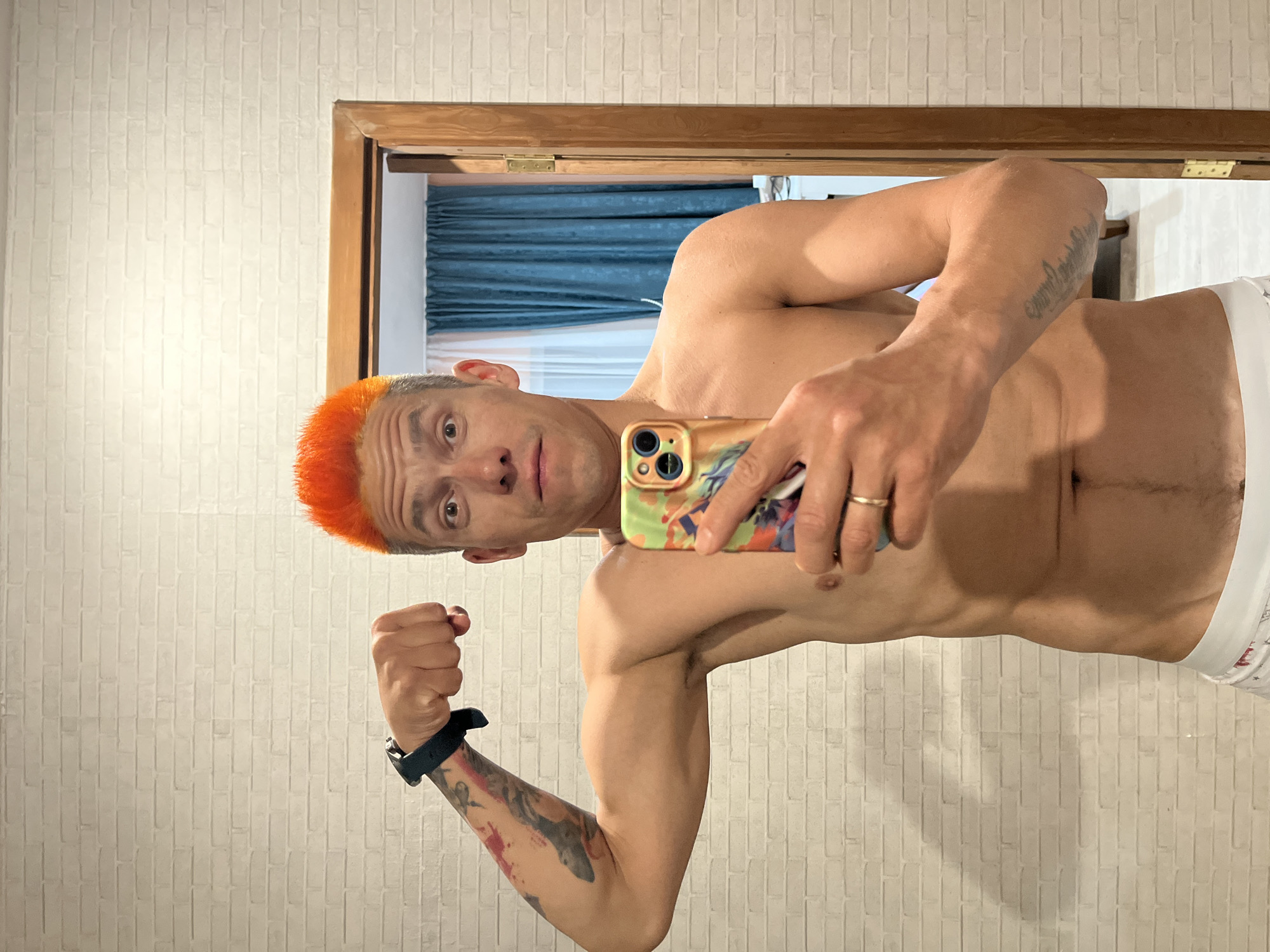 A shirtless man with orange hair is taking a selfie in front of a mirror. He is flexing his left arm, which has a black smartwatch and tattoos. He is holding a phone in his right hand, wearing a gold wedding band. The background includes a beige brick-patterned wall, a wooden door frame, and blue curtains visible through the doorway. The location is possibly in Chaikovsky, Russia.
