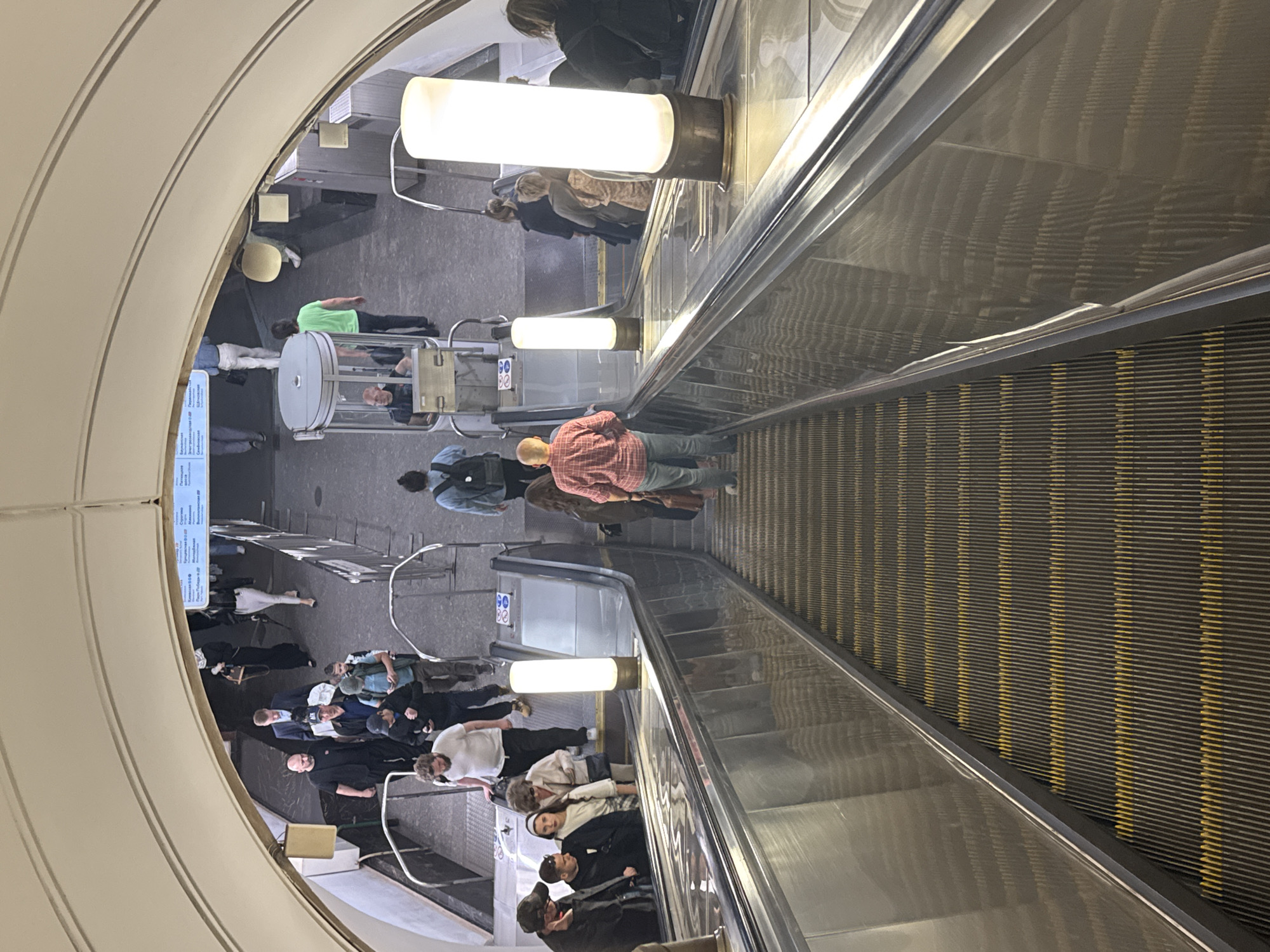 The image shows an overhead view of people on escalators in a Moscow Metro station. Several individuals are ascending and descending on parallel escalators. The escalators are made of metal and have yellow safety stripes. Tall, cylindrical light fixtures are positioned between the escalators. People are dressed in casual clothing, with some carrying bags or backpacks. Signs with Russian text are visible in the background, indicating this is a public transit station in Moscow. The architecture features a curved, light-colored ceiling with exposed beams.