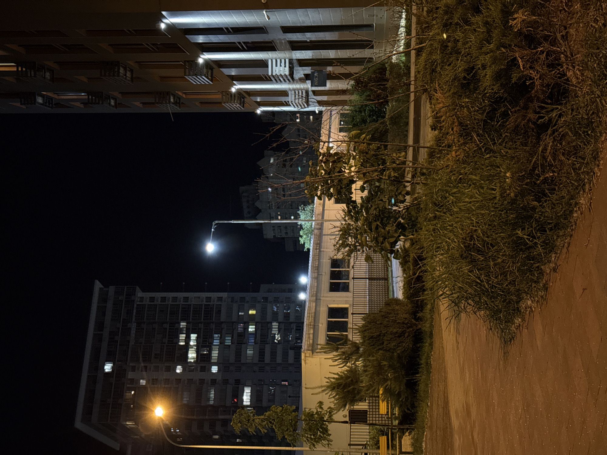 This media file captures a nocturnal urban landscape in Kyiv, Ukraine. The scene is dominated by several multi-story buildings, illuminated by artificial lights against a dark night sky.
In the foreground and along the right side, a paved path or ground area is visible, bordered by dense vegetation including bushes and trees with both lush foliage and bare branches.
On the left side of the frame, a prominent, large building with a distinctive angled facade features numerous lit windows, suggesting active occupancy. In the distance, towards the center, another tall, dark building is seen with several illuminated windows scattered across its structure. Part of a third building, possibly more modern, is visible in the upper right, displaying a structured facade with integrated lighting.
The environment is lit by a mix of streetlights and building lights. Several streetlights emit a warm, yellow glow, creating noticeable light flares, particularly in the lower left and central areas. Other lights, including those from building interiors and some streetlights, emit cooler white light.
A small sign displaying the letters "N.G.A" is faintly visible on the facade of a building in the upper right portion of the image. No people or specific activities are discernible within the scene, conveying a quiet, still atmosphere.