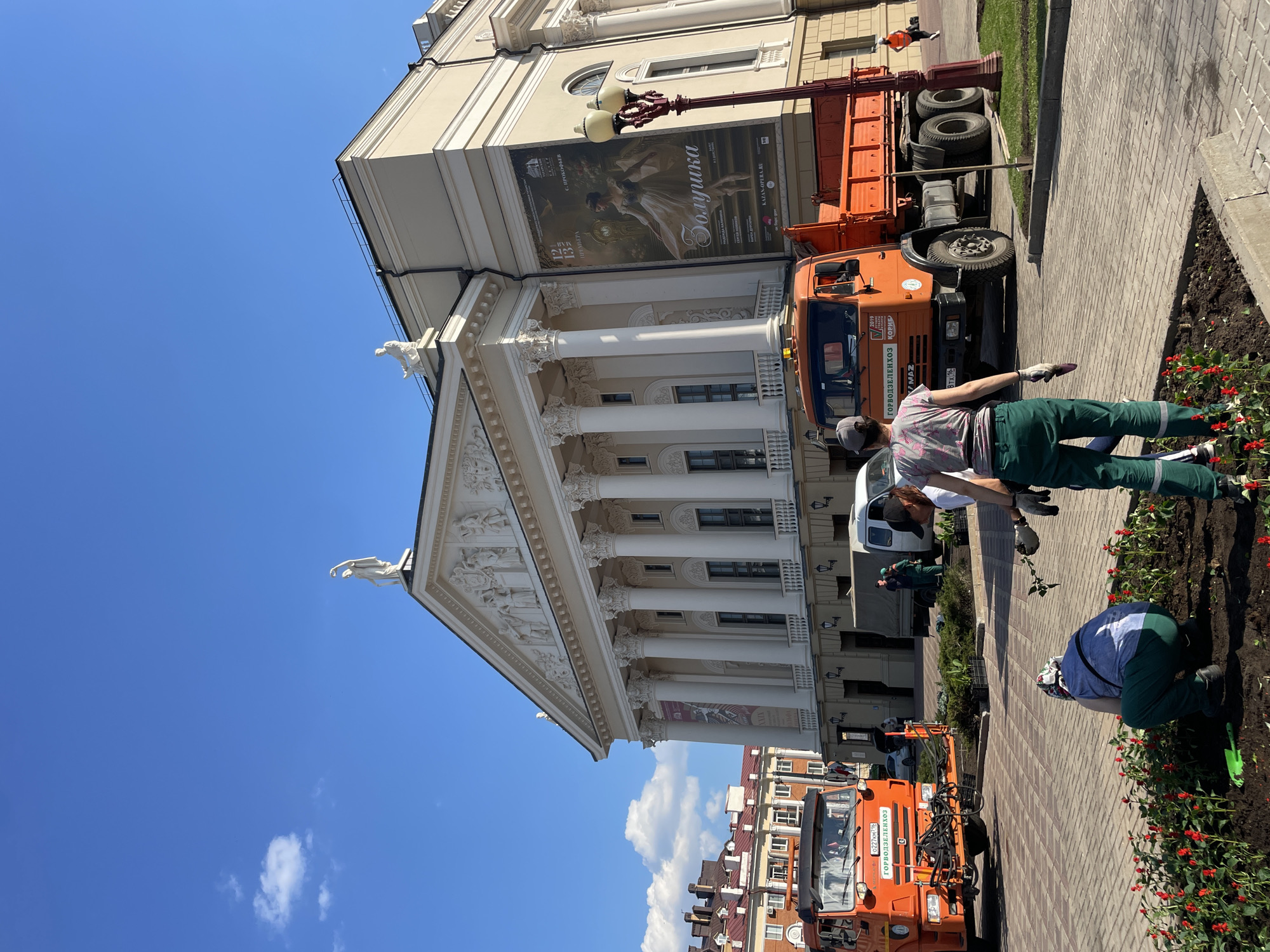 The image depicts a scene in Kazan, Russia, likely near the Musa Jalil Tatar Academic State Opera and Ballet Theatre. The theatre building is visible, featuring a classical façade with columns, statues, and a banner advertising the ballet "Zolushka" (Cinderella) by Sergei Prokofiev. Several workers wearing green uniforms are tending to flowerbeds along the sidewalk. One worker is kneeling and planting flowers. Two orange trucks belonging to the city landscaping company, "ГОРВОДЗЕЛЕНХОЗ" (Gorvodzelenkhoz), are parked nearby. The sky is blue with a few scattered clouds. The sidewalk is paved with rectangular bricks. The image captures a sunny day during landscaping activity near a prominent cultural landmark in Kazan.