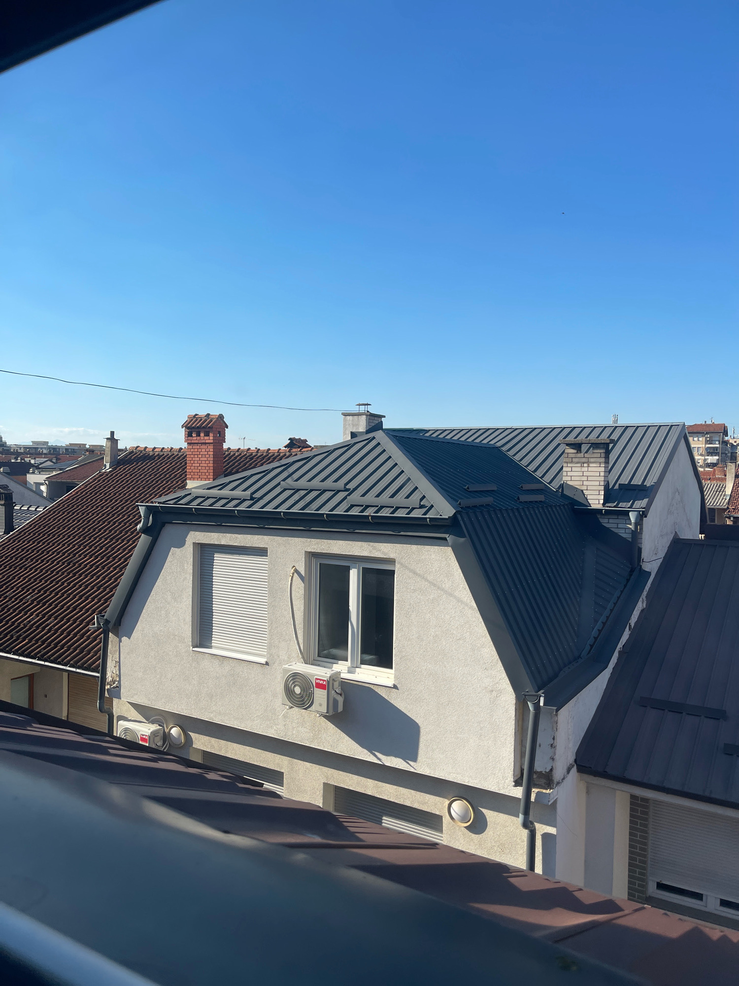The image shows a view of rooftops and buildings under a clear blue sky in the city of Prizren, Kosovo. In the foreground, a dark, angled surface, likely another rooftop or balcony railing, partially obscures the view.
The central focus is a white house with a dark, modern metal roof. The house has two windows facing forward, one with white horizontal blinds and another with clear glass. An outdoor air conditioning unit is mounted on the wall below the clear window. To the left of this house, a neighboring building features a reddish-brown tiled roof and a brick chimney with a decorative top. Further in the background, other buildings and rooftops are visible, some with tiled roofs and others with metal roofs, indicative of an urban or suburban setting. The sky is clear and bright blue, suggesting a sunny day. There are no people or animals visible in the image. The scene is static and appears to be a daytime landscape.