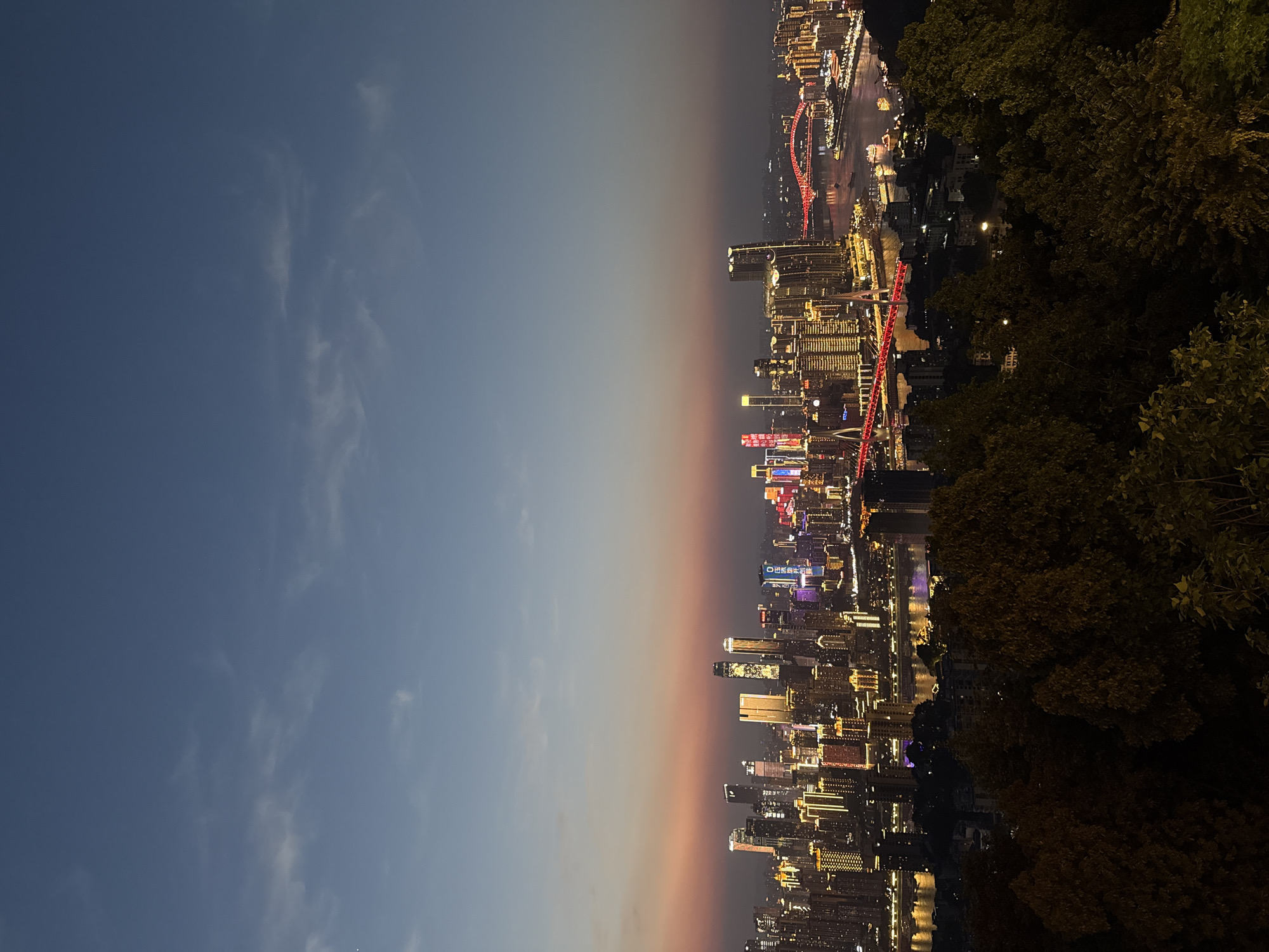 The image depicts a nighttime cityscape viewed from an elevated position, likely within Nan'an District, China. The right side of the frame is dominated by dark foliage from trees in the foreground. The city itself is brightly illuminated with varied lighting, including lights in the buildings, street lights, and decorative lights. A bridge, lit in red, spans a river that reflects city lights. The sky transitions from a light blue to a gradient of orange and grey, suggesting either dusk or dawn. The image is oriented with the horizon running vertically through the middle of the frame.