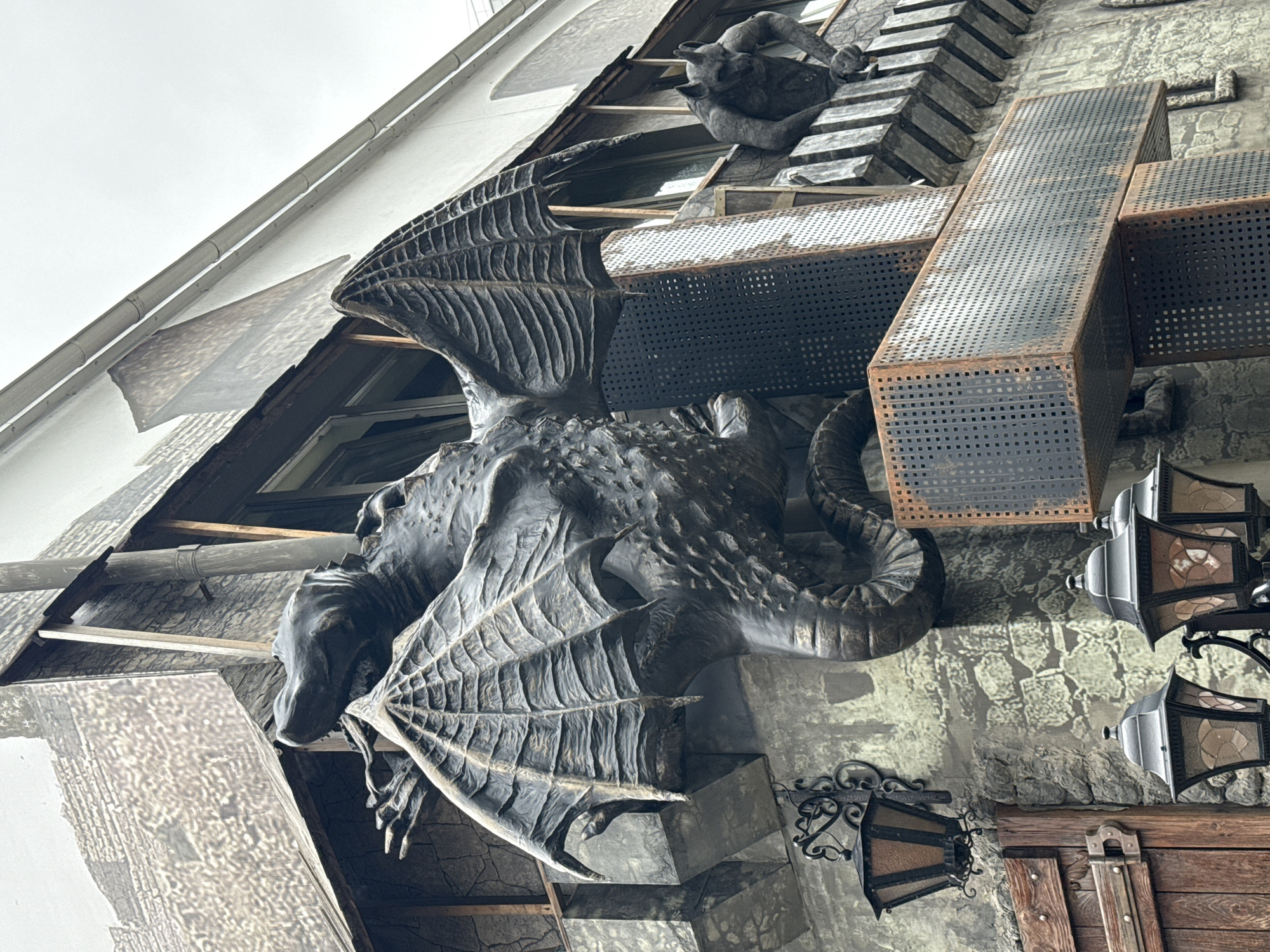 This media file shows a close-up, upward-angled view of a building facade adorned with a large, dark sculpture of a dragon. The dragon sculpture is highly detailed, with textured scales, large wings with visible bone structure and webbing, and a serpentine tail. It appears to be made of a dark, weathered material, possibly metal or cast stone.
The dragon is mounted on the side of a building, which features a mix of architectural styles and materials. To the left of the dragon, a section of the building is covered in a white material with a subtle textured pattern, possibly a tarp or decorative element. Beneath this, wooden beams and what appears to be a window or opening are visible.
To the right of the dragon, the building's wall has a rough, stone-like texture. Several metal architectural elements are attached to the facade, including a large, cross-shaped structure made of perforated metal plates. Two ornate, dark metal lanterns with glass panels are also visible, hanging from the wall. Above the dragon, two smaller, dark sculptures resembling gargoyles or smaller creatures are perched on a tiered structure.
The scene appears to be an outdoor setting, with the sky visible in the upper left quadrant of the image, suggesting an overcast or bright day. There are no people visible. The overall impression is one of fantastical, gothic-inspired architecture. The specific location is identified as Perm, Russia. No visible text is present in the image.