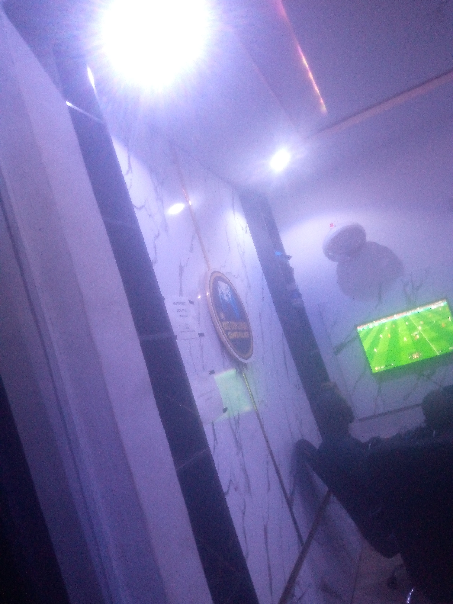 The image depicts an indoor scene, likely a gaming or entertainment lounge, located in Jalingo, Nigeria. A powerful, overexposed white light source in the upper left foreground dominates the frame, creating a significant starburst flare and casting a purple-blue ambient light across the entire space. Other smaller, recessed light fixtures are visible along the ceiling.
The main wall in the background is covered with a white marble-patterned surface featuring dark grey veining. Mounted on this wall, slightly left of center, is a circular, gold-rimmed plaque. The text on the plaque is partially legible, appearing to read "HORIZON CLUB" or similar at the top and "GAMING PALACE" or "GAME PALACE" at the bottom. Several white paper notices or flyers are taped to the wall near the plaque, one displaying numerical values like "300" and "200," possibly indicating pricing or instructions.
To the right of the plaque, a flat-screen television is mounted on the wall, actively displaying a football (soccer) video game with a vibrant green pitch and visible player avatars. A score or game interface element is visible in the lower right portion of the screen.
In the mid-right, a white, circular fixture, possibly a decorative light shade or a small ceiling fan, hangs from the ceiling. In the lower right quadrant of the image, two dark, indistinct human silhouettes are visible, seated and facing the television, suggesting they are observing or playing the game. One figure is slightly more defined, showing a head and shoulders. The flooring is not discernible, and the space appears to be dimly lit overall, contrasting with the bright artificial light sources and the television screen.