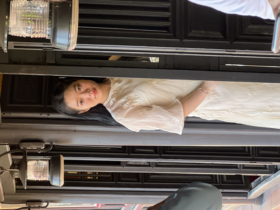 A young East Asian woman is partially visible, positioned slightly off-center to the right, looking directly at the viewer with a neutral expression. She has long dark hair and is wearing a light-colored, possibly cream or off-white, long dress adorned with embroidered floral patterns on the bodice and elbow-length, flared sleeves. A silver-colored bracelet is visible on her left wrist. She stands partially obscured by a dark, vertical wooden door frame or panel. In the background, dark wooden paneled doors or shutters with traditional detailing are visible. Two dark-colored, wall-mounted lanterns with clear, ribbed glass shades hang in the upper corners of the frame. To the lower left, the shoulder and back of an individual in a light blue garment are partially visible. To the lower right, a hand holding a mobile phone is partially visible, belonging to an individual wearing a white shirt. The scene is brightly lit, indicating daylight. This setting is observed in Nha Trang, Vietnam.