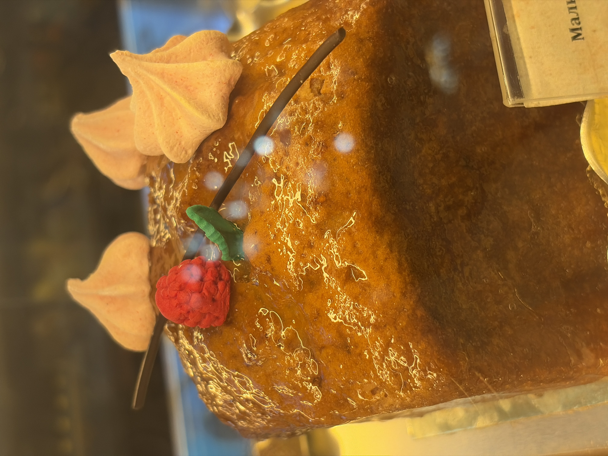 The image shows a close-up of a rectangular cake with a glossy, amber-colored glaze. The cake is decorated with two pale orange meringue kisses, a single red raspberry, and a thin dark stick that appears to be a skewer or twig. A small green leaf is also visible, likely part of the decoration. In the lower right corner, a label with Russian text, partially obscured, is visible. The lighting suggests the cake is displayed in a bakery or pastry shop, possibly behind glass. The reflection of lights on the glaze indicates an indoor environment. The label's visible text, "Мали" (Mali), likely refers to "raspberry" in Russian, hinting at the cake's flavor. The presence of such a display is common in St. Petersburg bakeries, particularly in areas like Nevsky Prospekt or around major historical sites where tourist-oriented shops are prevalent. However, without further context or landmarks, the precise location in Saint Petersburg cannot be determined.
