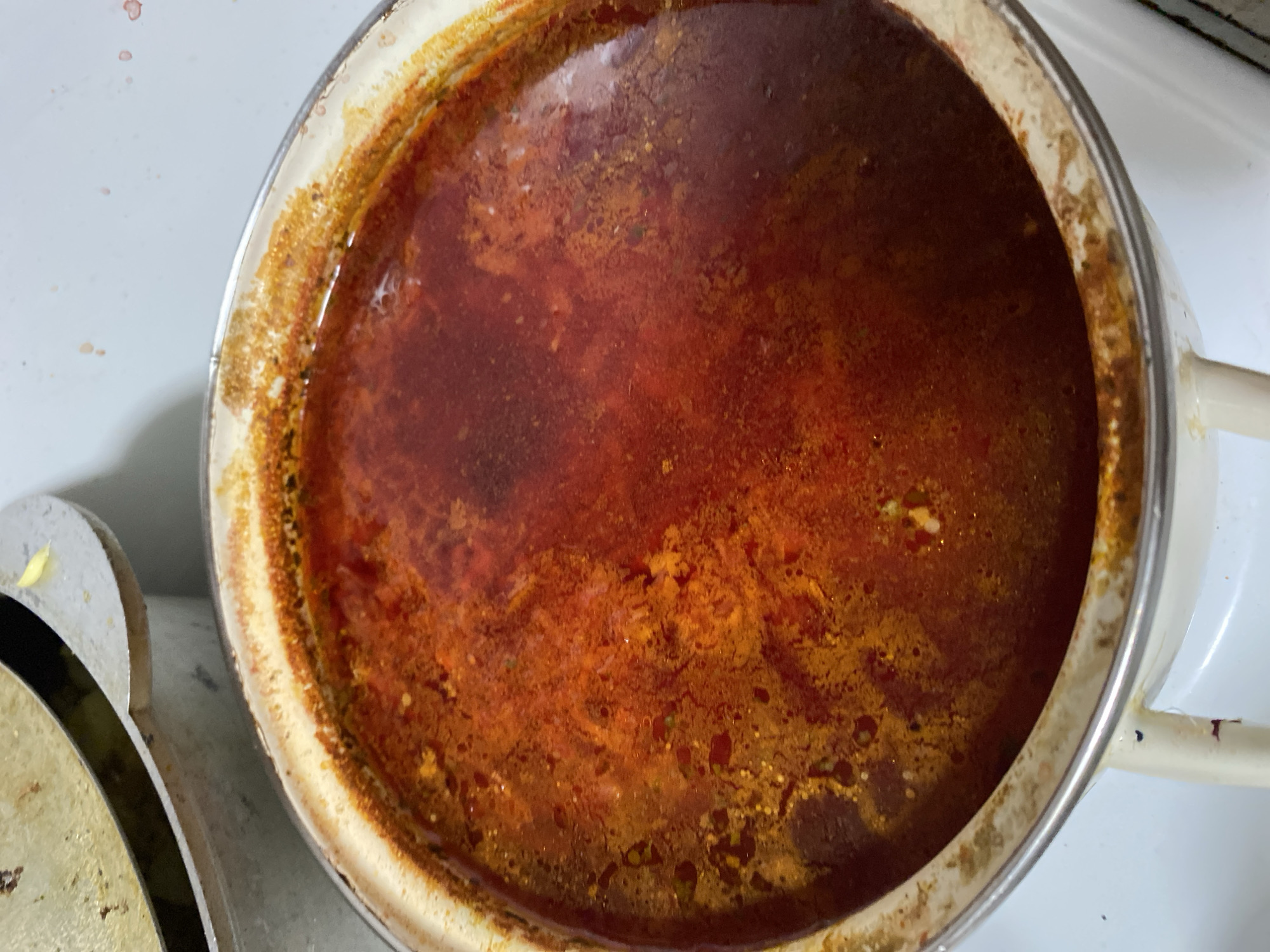 A close-up, overhead view shows a pot of simmering red stew, likely borscht, in a kitchen in Chornomorsk, Ukraine. The stew is a deep reddish-brown with visible pieces of vegetables and fat. The pot is white with a metallic rim and handle, and it sits on a white stovetop. A second pot with a metallic lid is partially visible in the upper left corner of the image. The scene appears to be domestic, with no people or animals present.