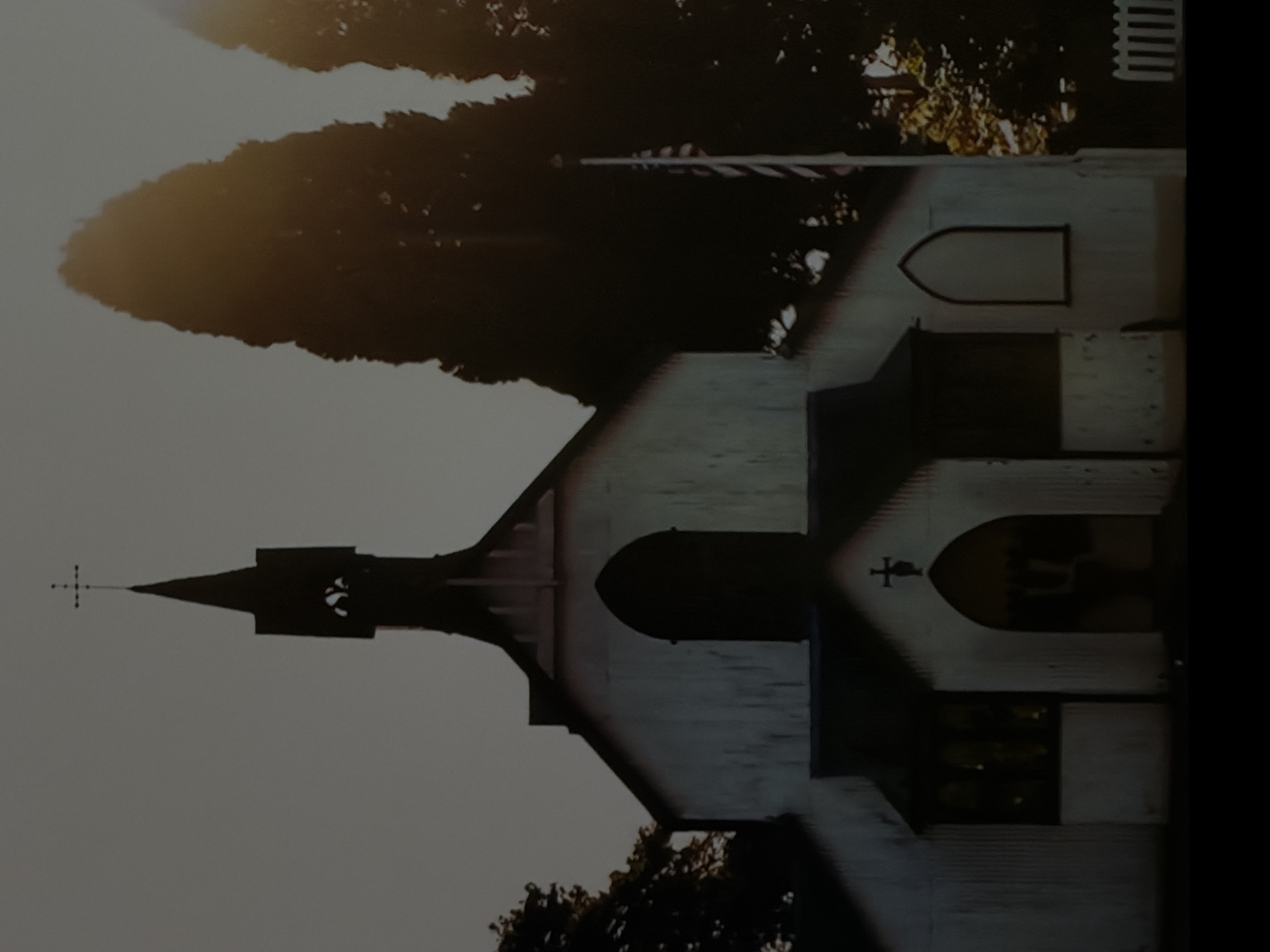 The image displays a serene view of a white or light-colored church under a soft, light grey sky. The lighting, characterized by warm, golden hues and long shadows cast across the building, suggests either early morning or late afternoon.
The primary subject is the church, which features a tall, dark spire topped with a cross. Another, smaller cross is visible above an arched entryway on the side of the main building. Several arched windows, appearing dark or reflective, are set into the church's walls. The architecture shows distinct lines and possibly wooden siding or stucco, creating textural depth.
Surrounding the church are several trees, with one particularly large, dark-foliaged tree standing prominently behind it, catching the golden light on its leaves. A flagpole is visible to the right of the church, from which an American flag is flying. A section of a white picket fence is also partially visible further to the right.
The scene is static, with no human figures or discernible activity taking place. The overall impression is one of peace and quiet. There is no visible text within the image.