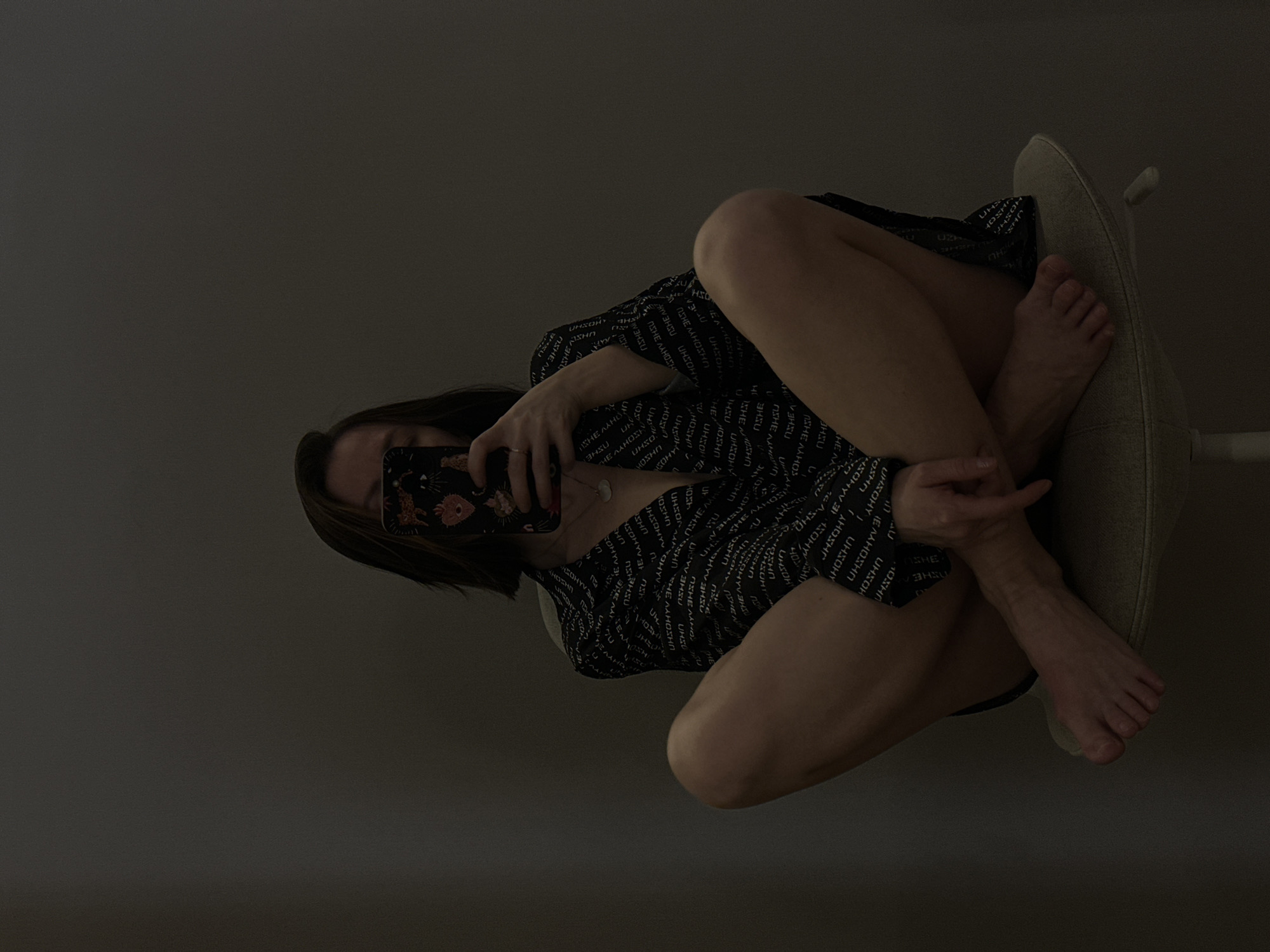 A person is sitting in a chair taking a selfie in a dimly lit room. The person is angled towards the camera, knees up, and wearing a patterned, long-sleeved shirt. The individual holds a phone in front of their face. The room appears to have minimal lighting and a plain wall in the background. The location is not identifiable.