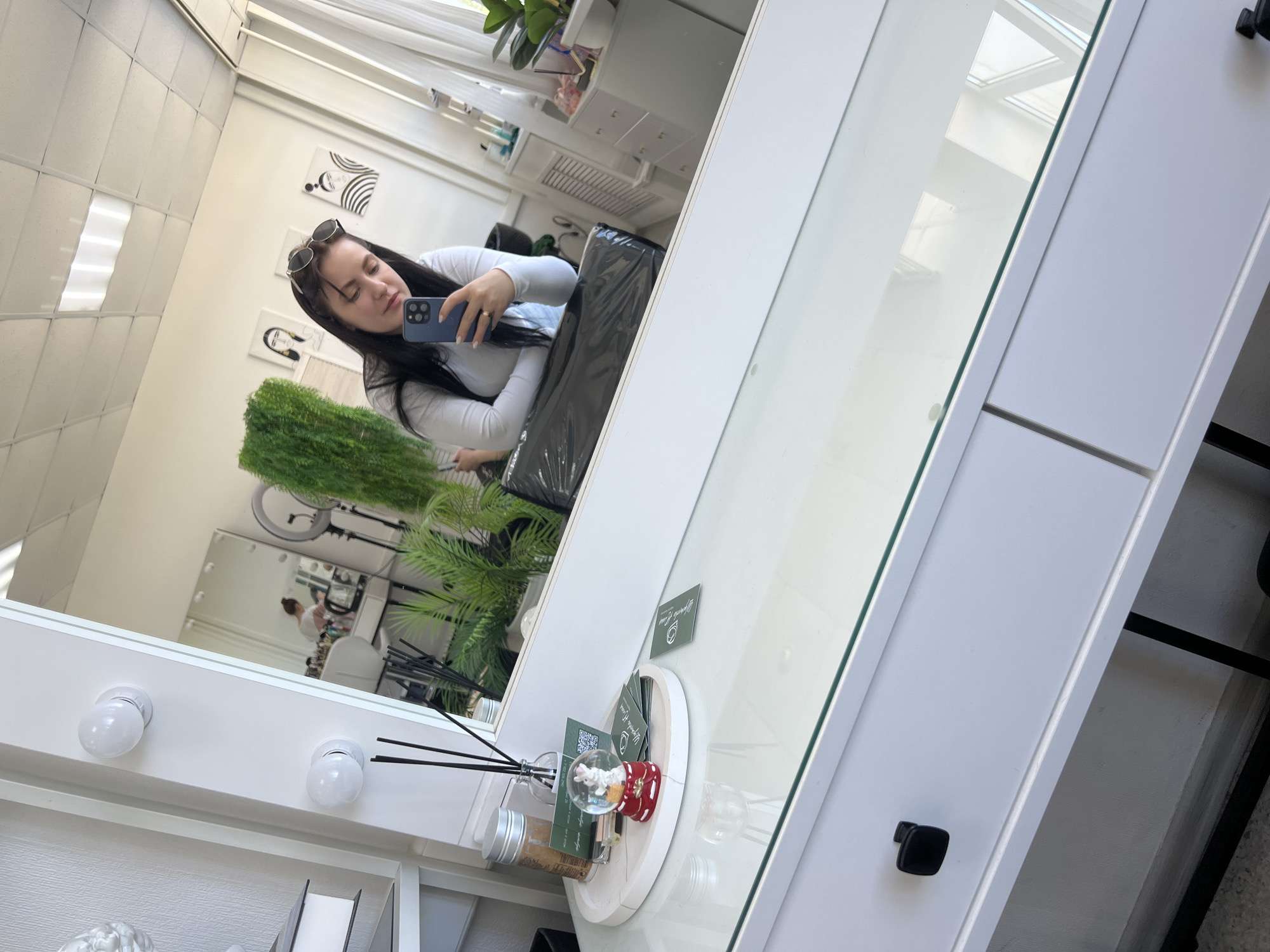 A woman in a white top is taking a selfie in a mirror. The location appears to be inside a beauty salon, with white walls, bright lighting, and various salon-related items visible. The image is taken from a low angle, looking upwards towards the mirror. The location is assumed to be in the city of Chaikovsky, Russia.
