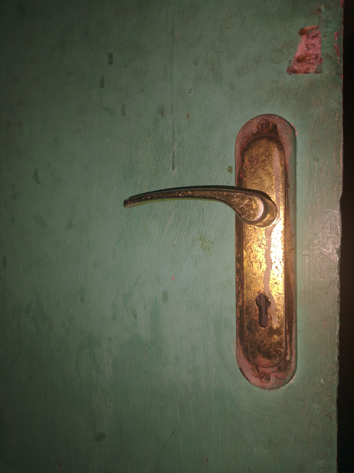 The image shows a close-up of a tarnished brass doorknob and lock mechanism mounted on a green wooden door. The door surface exhibits visible scratches and some peeling paint in the upper right corner. The doorknob is a curved lever handle, and below it is a keyhole. The overall impression is of an older, somewhat worn door. The location in Mukapayung, Indonesia, is not visually discernible from the image itself.