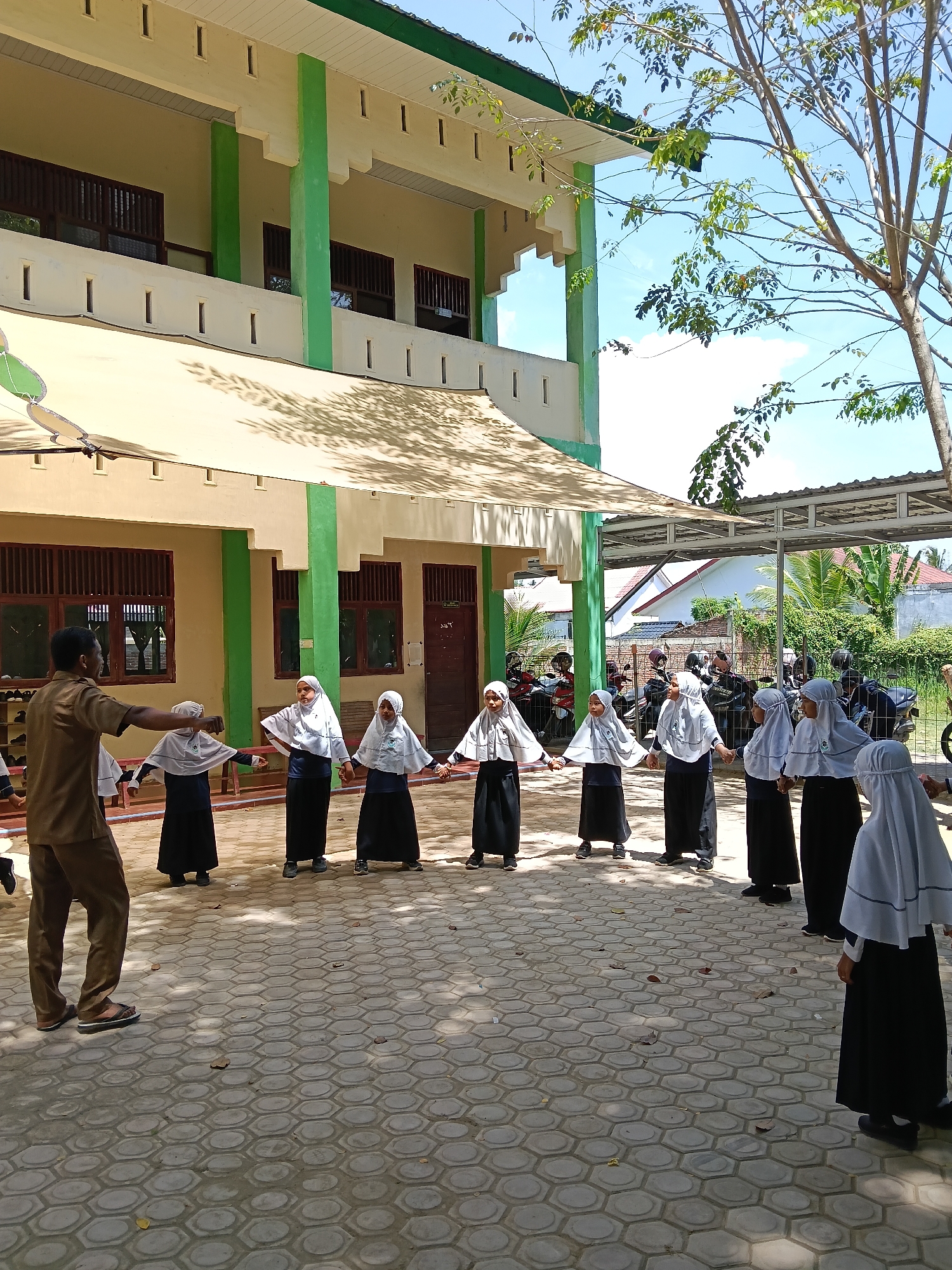 A group of nine children, all female, are arranged in a circle on a paved hexagonal-tiled courtyard. They are wearing black skirts, dark tops, white hijabs, and shoes. They are holding hands and appear to be participating in an activity or game led by a man standing to the left of the frame. The man is wearing a brown shirt and brown pants, and he has his arms outstretched.
Behind the group is a two-story building with a green and beige color scheme. The building has windows with wooden slats and a covered walkway. In the background, behind a metal structure, are trees and a glimpse of a white house. Motorbikes and people are also visible in the background. The sky is blue with some clouds, and there is a tree with sparse foliage in the upper right corner of the frame. The shadows cast across the courtyard suggest it is a sunny day. The scene is located in Lhokseumawe, Indonesia.