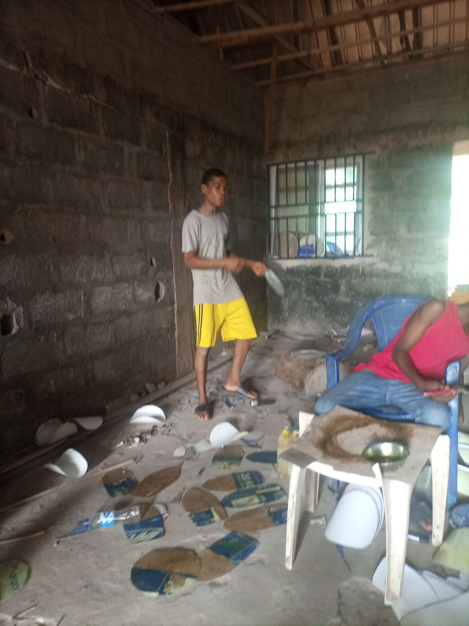 This image depicts an indoor scene in Nkpor, Nigeria, likely a workshop or an unfinished building. Two young men are present. One stands in the center of the room, wearing a grey t-shirt and bright yellow shorts, holding a flat object, possibly a piece of wood or plastic, in his hands. He appears to be in motion, perhaps engaged in a craft or activity. The second individual is seated on a blue plastic chair to the right, wearing a red tank top and blue jeans, and is looking down at something in his hands, possibly a mobile phone.
The setting is rustic and appears to be under construction or in a state of disarray. The walls are made of cinder blocks, and the ceiling shows exposed wooden beams and roofing material. A window with metal bars is visible in the background. The floor is a concrete surface scattered with various objects. Among these are white, curved items that resemble shoe soles or components, along with irregularly shaped pieces of material that look like cut leather or fabric, possibly for shoe making. A plastic bottle of water and a small metal bowl on a makeshift wooden table are also visible.
The lighting suggests it is daytime, with natural light coming from the window. There are no obvious indicators of the weather. The overall atmosphere seems to be one of work or preparation, with the scattered materials hinting at a craft or manufacturing process. No specific text is clearly legible in the image.