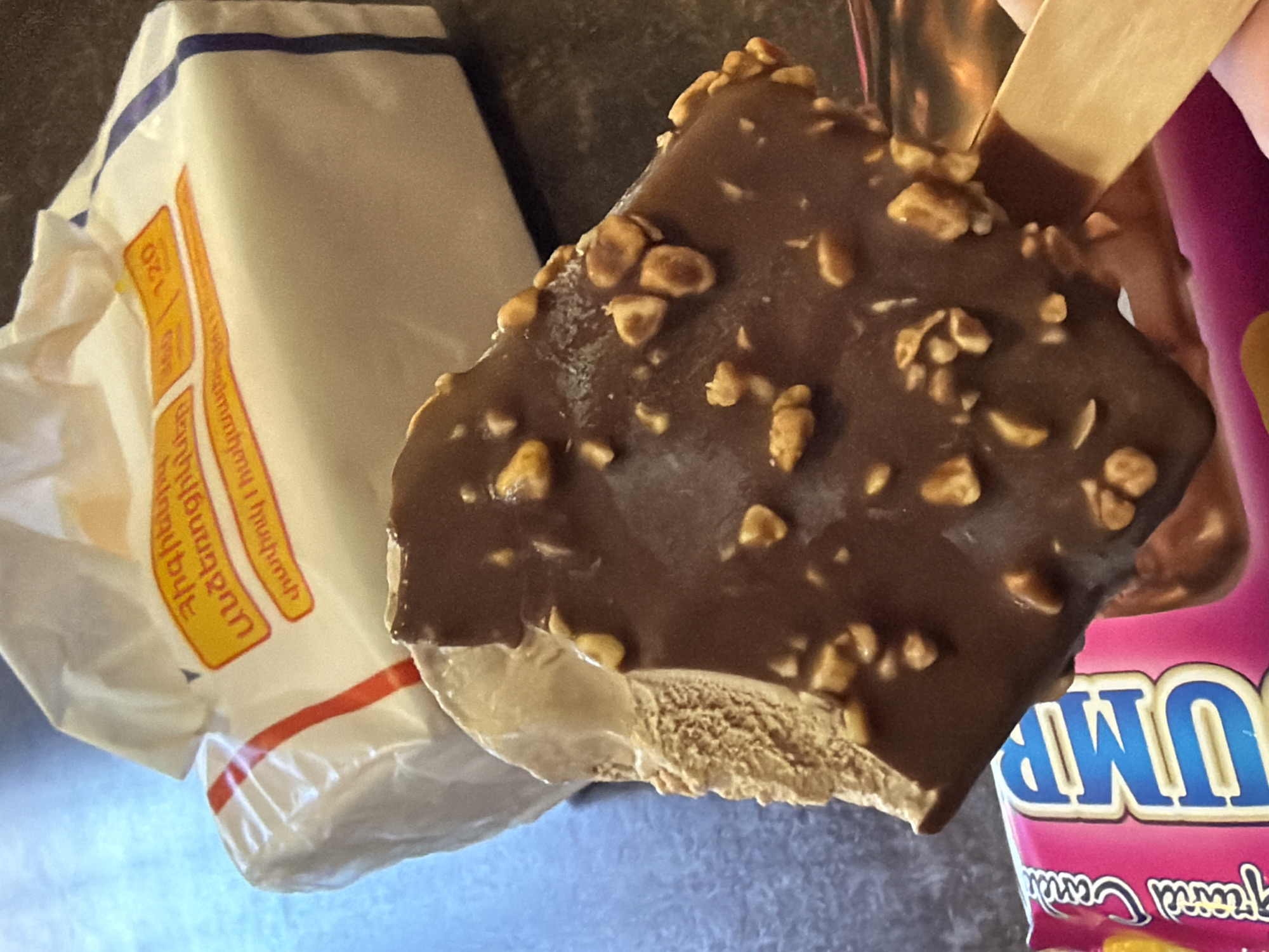 The image depicts a partially eaten ice cream bar with a chocolate coating and nut pieces, held by a wooden stick. Adjacent to the ice cream bar is a white and red wrapper. Part of another wrapper, possibly the original packaging of the ice cream, is also visible with the word "JUMB" written in large blue letters on a pink background. The wrappers have Armenian writing on them. No people are visible. No specific details indicate the exact location in Merdzavan, Armenia, only that the wrappers have Armenian writing.