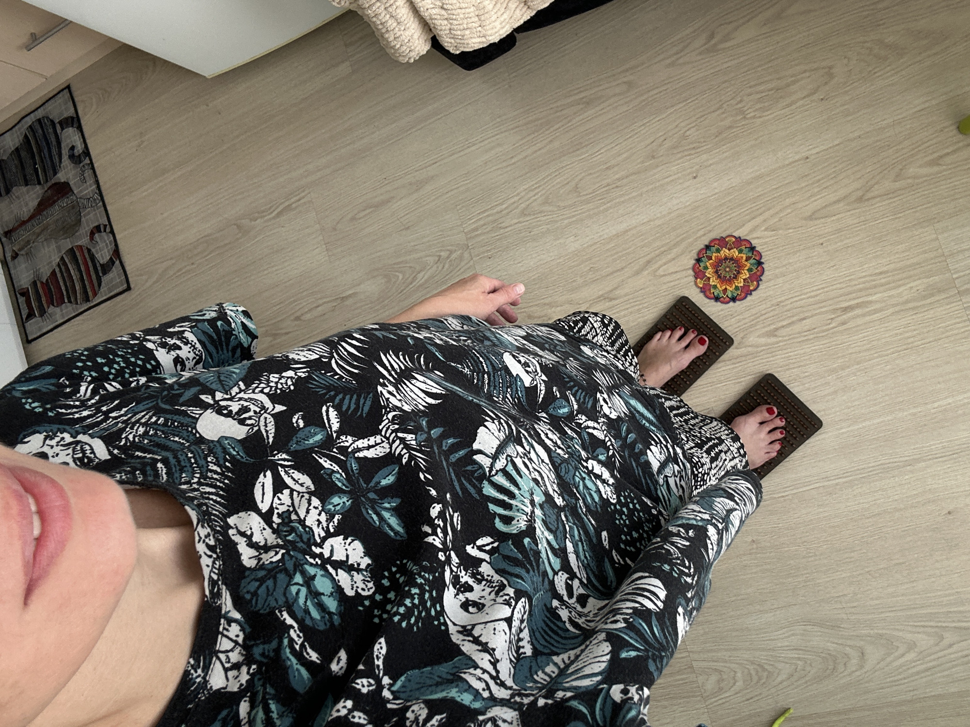 A person stands on a light-colored wooden floor in a room, wearing a black and teal patterned long-sleeved dress. Their feet are bare except for red nail polish, and they are standing on two dark brown massage mats. To the right of the person, there is a small, colorful mandala-patterned coaster. In the background, there is a rug with a pattern of three cats, and a portion of a light-colored cabinet. To the right of the cabinet, a stack of folded, textured cream-colored blankets is visible. The scene does not provide specific details to identify the location as Kemerovo, Russia.