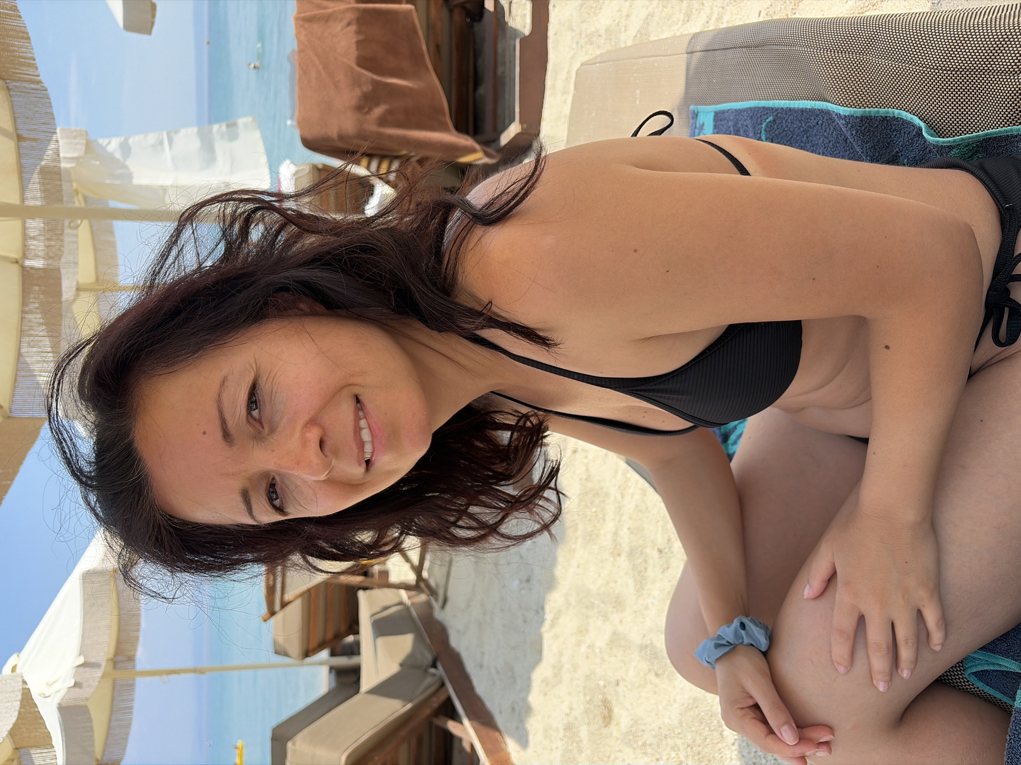 A woman with dark, wavy hair and tanned skin is sitting on a lounge chair on a sandy beach. She is wearing a black bikini and a light blue scrunchie on her left wrist. Her hands are resting on her lap. In the background, the turquoise sea is visible under a clear blue sky, with beach umbrellas and lounge chairs suggesting a resort setting in Pallini Municipal Unit, Greece. The overall scene depicts a relaxed day at the beach.