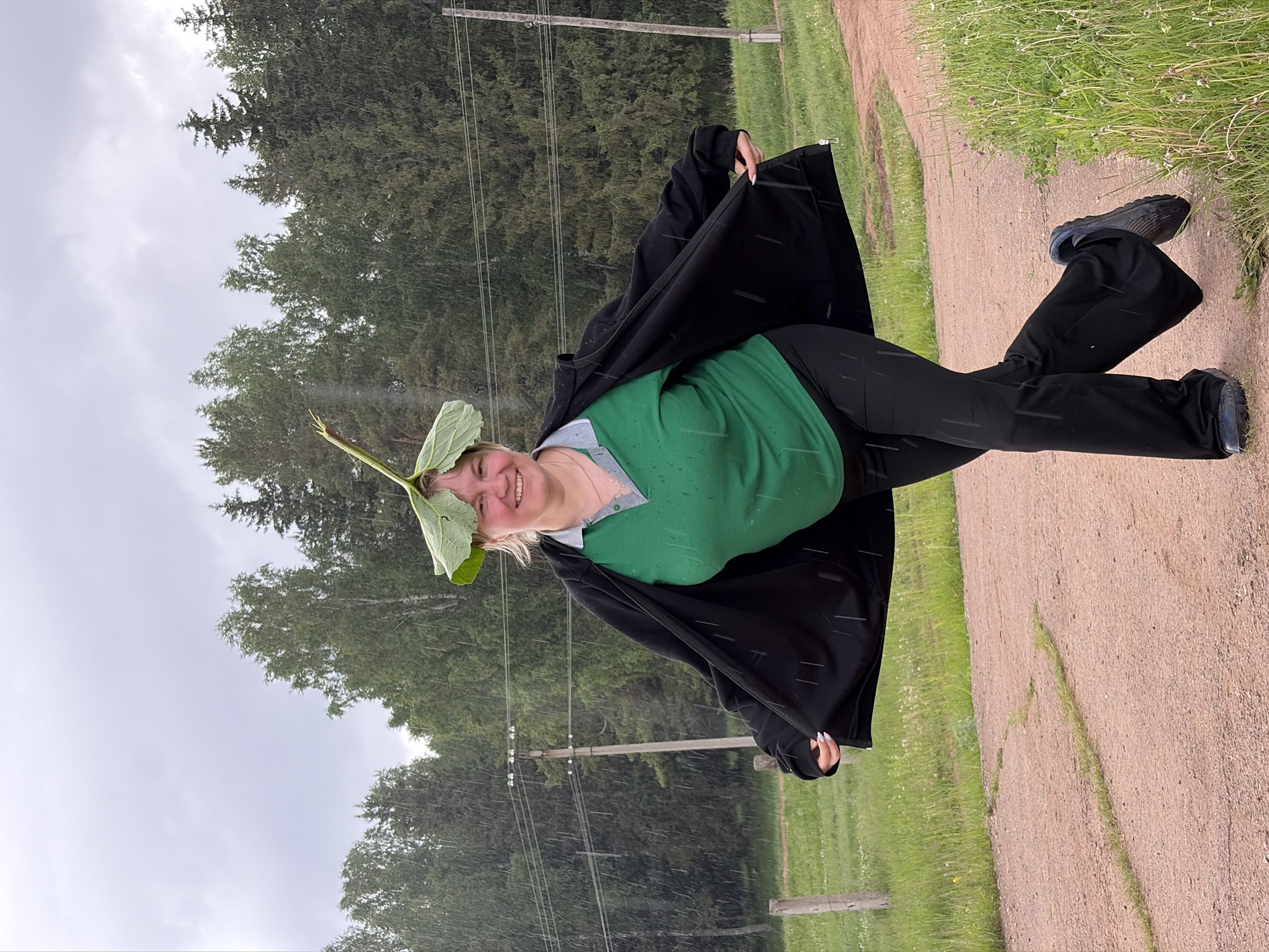 A woman stands on a dirt path under a rainy sky in what appears to be a rural area of Nizhnii Vishur, Russia. She is wearing a green shirt, black pants, and a black jacket. A large green leaf rests on her head. She is smiling. Trees and power lines are visible in the background. The ground is damp, and rain streaks are visible throughout the image.