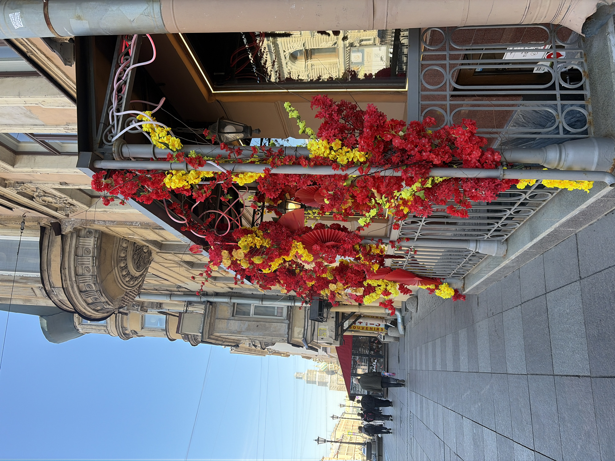 This image depicts a vibrant street scene in Saint Petersburg, Russia, under a clear blue sky. The perspective looks down a pedestrian walkway, flanked by buildings on both sides.
The most prominent feature is an elaborate overhead decoration installed along a building on the right. This festive display consists of numerous artificial red and bright yellow flowers, resembling blossoms, intertwined with swirling pink neon-like lights and complemented by several red fan-shaped ornaments. This design evokes an East Asian aesthetic, suggesting the presence of a themed establishment, such as a restaurant or shop, below.
The building on the right, adorned with these decorations, features a dark facade with a large reflective window that mirrors the classical architecture of the opposite side of the street. Below the decorations, a grey metal grate with a pattern of circular and oval shapes is visible. On the left, buildings exhibit ornate classical European architectural elements, including decorative cornices, window frames, and balconies.
Several people are seen walking casually on the wide, dark grey tiled pavement in the mid-ground, appearing as indistinct figures. Further down the street, a red awning marks a storefront, and a partially visible sign above it reads "SOUVENIRS." The bright, clear sky suggests it is daytime, likely mid-morning or afternoon, with pleasant weather. The overall scene portrays a lively urban commercial area, enhanced by the colorful floral canopy.