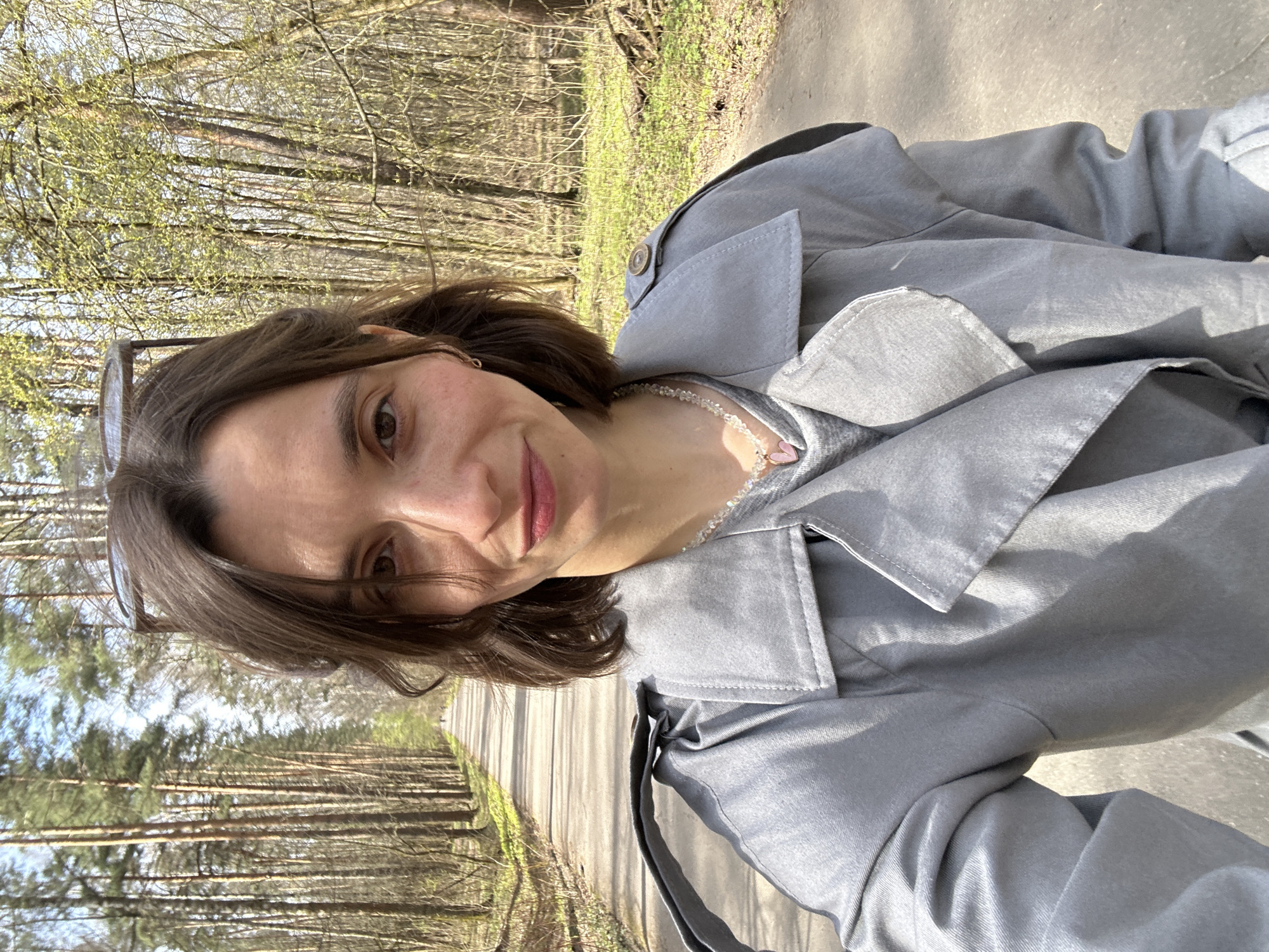A selfie taken outdoors on a tree-lined path. A young woman with short brown hair wears a gray trench coat and a delicate necklace. Her sunglasses are pushed up on her head. The path stretches into the distance, flanked by trees and dappled sunlight. The location appears to be a park or forest area. Based on the prevalent coniferous trees and the general aesthetic, the image could potentially be taken within a park in Moscow, Russia, though this cannot be determined with certainty.