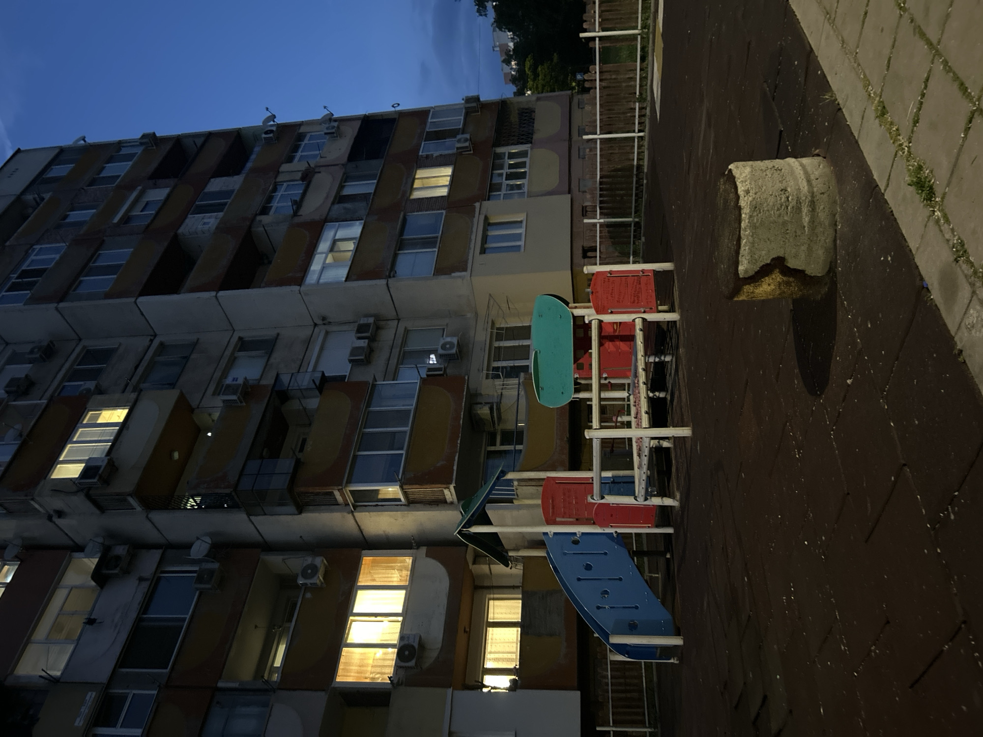 The image captures an urban scene at dusk in an unknown city. Dominating the left side of the frame is a tall, aging apartment building with a repetitive pattern of jutting balconies and windows, some of which are lit from within, casting a warm glow. Air conditioning units are visible on the exterior walls of the building. To the right of the building is a playground area with colorful play structures in shades of red, blue, and green, contrasting with the muted tones of the surrounding environment. A cylindrical concrete structure stands to the right, possibly serving as a barrier or seating. The ground is covered with dark rubberized flooring. A metal fence borders the playground area. The sky above transitions from blue to gray, indicating the fading daylight. There is no human presence visible in the image.
