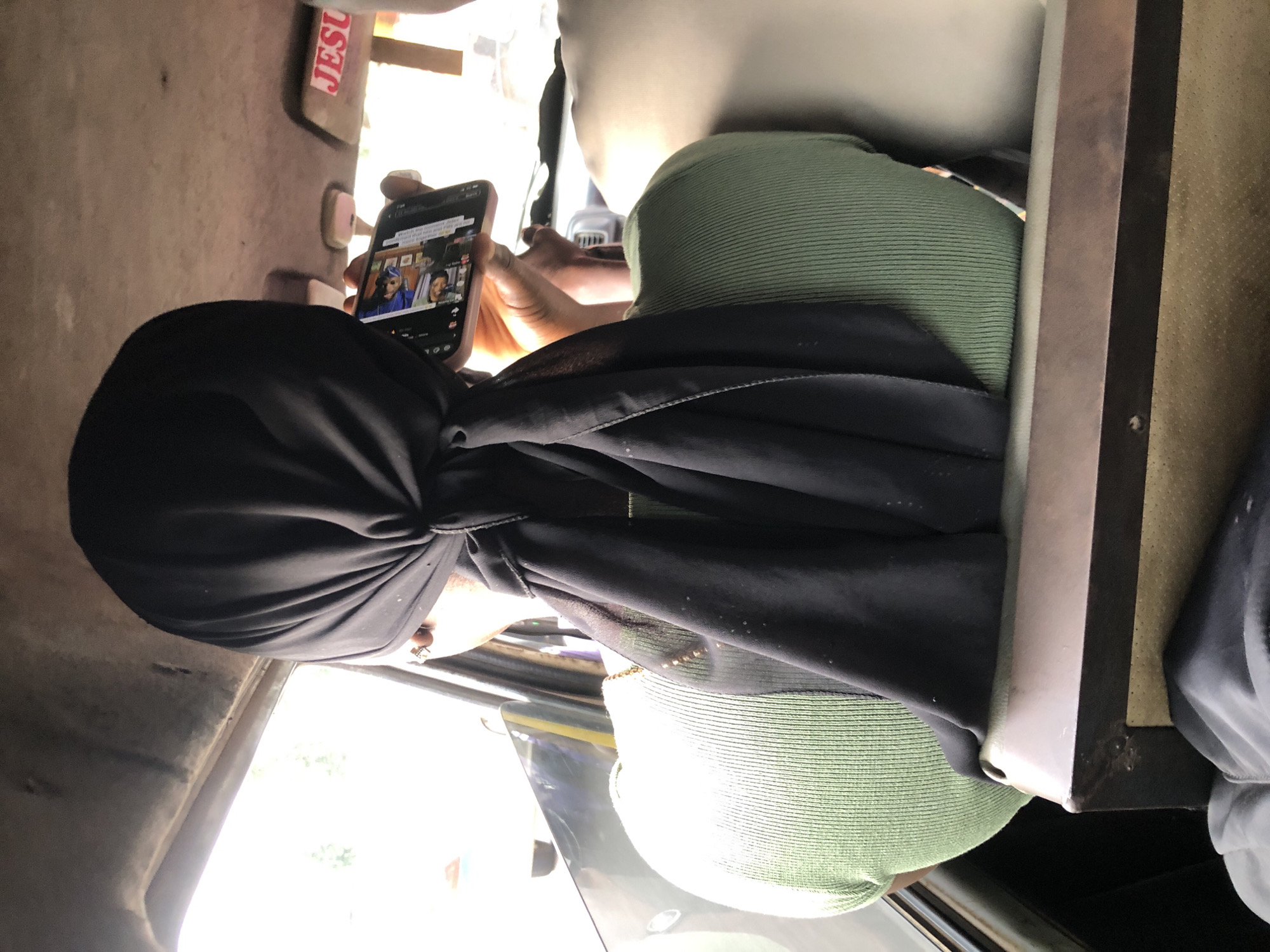 A rear view of a young woman wearing a black hijab and a light green ribbed shirt sits in the passenger seat of a vehicle. She holds a smartphone in her right hand, looking at the screen. The interior of the vehicle is visible, including the ceiling with a sun visor that has the word "JESU" partially visible on it, and seats in the background. Outside the vehicle, trees and a hazy sky are visible through the window. The scene appears to be in Shangisha, Nigeria.
