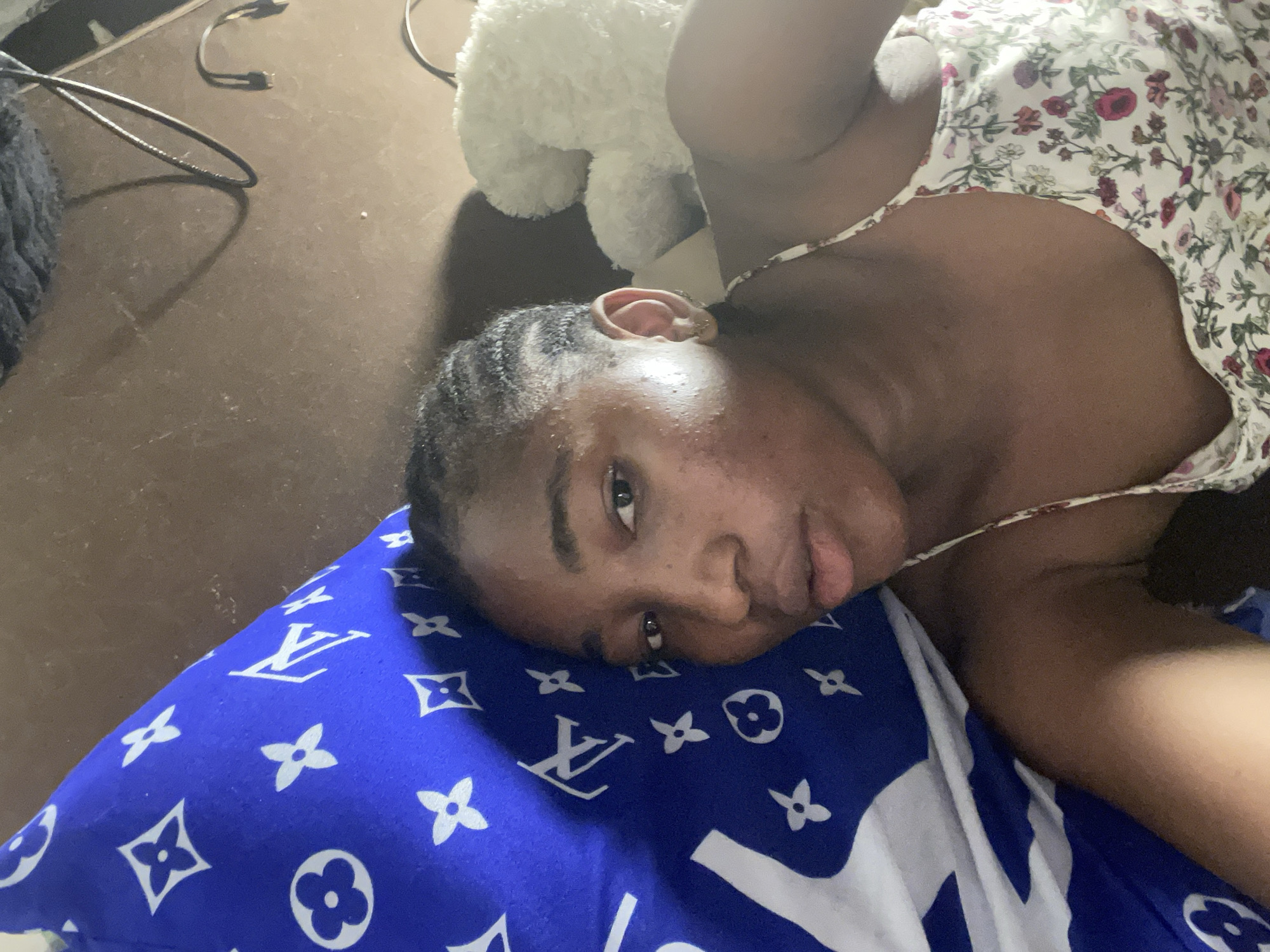 A person lies on a blue pillow with white patterns, dressed in a floral patterned top. A stuffed animal is near their head. The background includes a brown surface and some cables. There are no elements in the photo to suggest this image was taken in Lokoja, Nigeria.