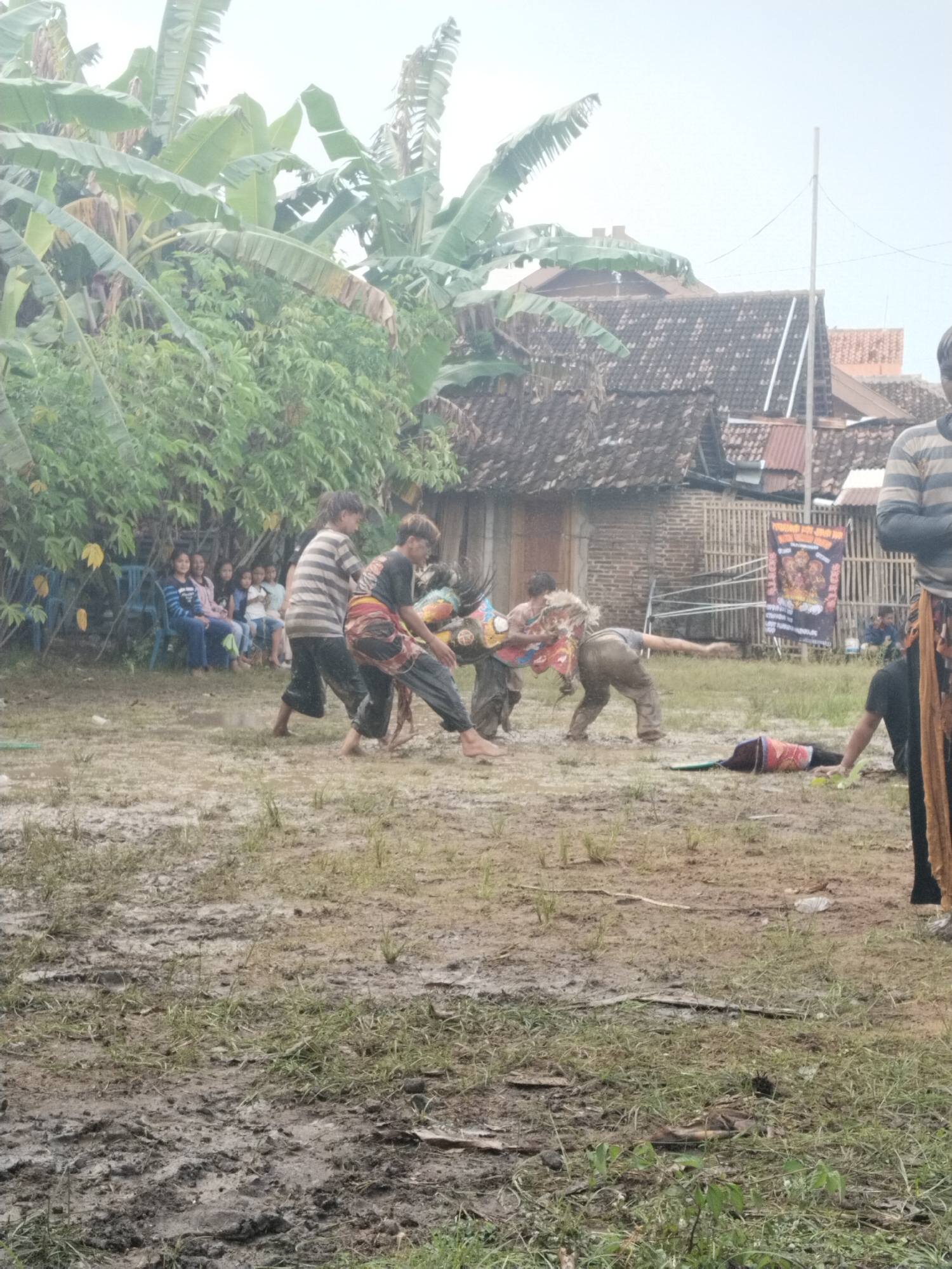 The image depicts a group of people engaged in what appears to be a traditional performance or game, possibly related to the local culture. The location is a muddy, open field, possibly after a rain, with some grass. In the background are simple houses, a banner with images and text, and some vegetation, including what appears to be banana trees.
Several individuals are in the center of the image, actively interacting. They are dressed in various clothes, some with traditional patterns or accessories. Some are barefoot. Their postures and movements suggest a physically active performance. A group of children or young people sit on chairs watching the performance.
The presence of the banner and the traditional attire suggest this is a local or cultural event. The muddy field and simple buildings indicate a rural or less developed area.