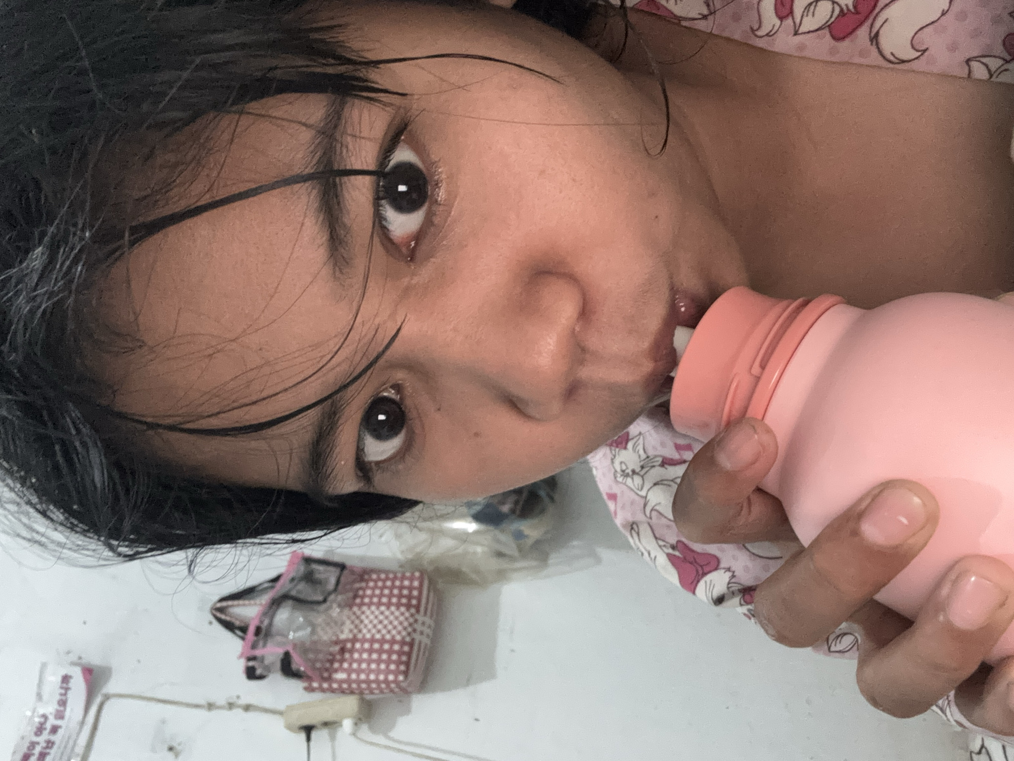 A close-up, low-angle, indoor selfie shows a person with dark, wet hair drinking from a pink bottle with a light pink lid. The person's face is centrally framed, with their eyes wide open and looking directly at the camera. Their lips are slightly parted as they drink. They are wearing a pink garment with a white pattern of what appears to be cartoon cats. The background is a plain, light-colored wall with some items visible on it: a white electrical outlet with a cord attached, a pink and white woven bag hanging on a hook, and a piece of paper with writing. The overall lighting is soft and somewhat dim. The scene appears to be in a residential setting in North Jakarta, Indonesia.