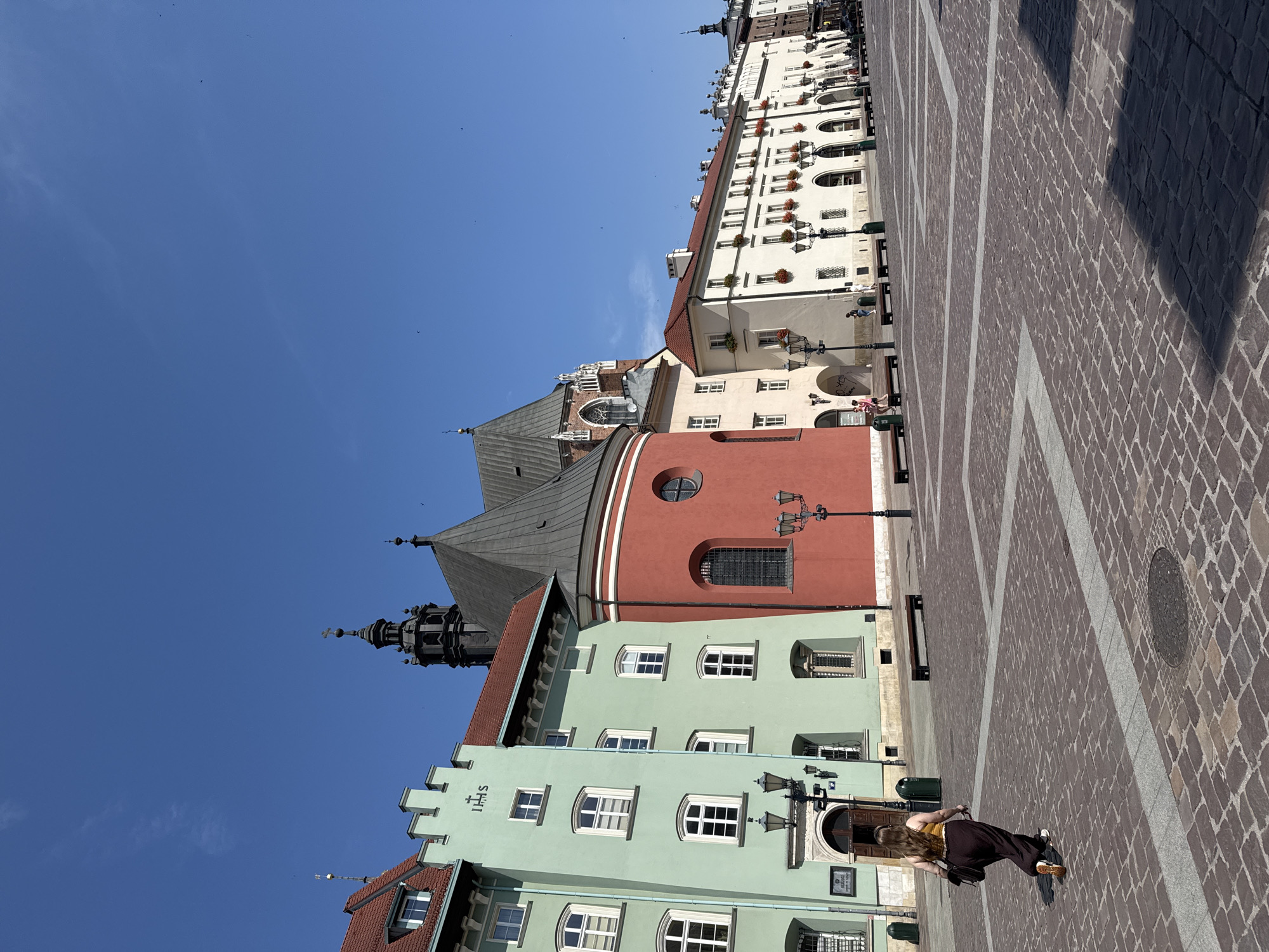 This is a medium shot, eye-level, outdoor, vertical photograph of the Main Market Square in Krakow, Poland, taken on a clear, sunny day. The square is paved with cobblestones, with lighter grey stone borders forming geometric patterns. In the foreground, a young woman with long, brown hair walks away from the camera towards the left side of the frame. She is wearing a yellow tank top and dark, flowing pants, and carrying a brown bag over her right shoulder. Her feet are bare, and she is wearing white sneakers.
Behind her, a row of colorful buildings lines the square. The leftmost building is pale green with red trim and a red roof, featuring multiple white-framed windows. The building next to it is a bright red cylindrical structure with a dark grey conical roof, accented by a Gothic architectural style spire in the background. Further to the right, a long, white building with an arched arcade runs along the square, adorned with numerous windows and flower boxes containing red flowers. Several black, ornate lampposts are visible throughout the scene, casting shadows on the cobblestone pavement. A few people are scattered throughout the square, some sitting on benches, while others walk. The sky is a vibrant, cloudless blue.