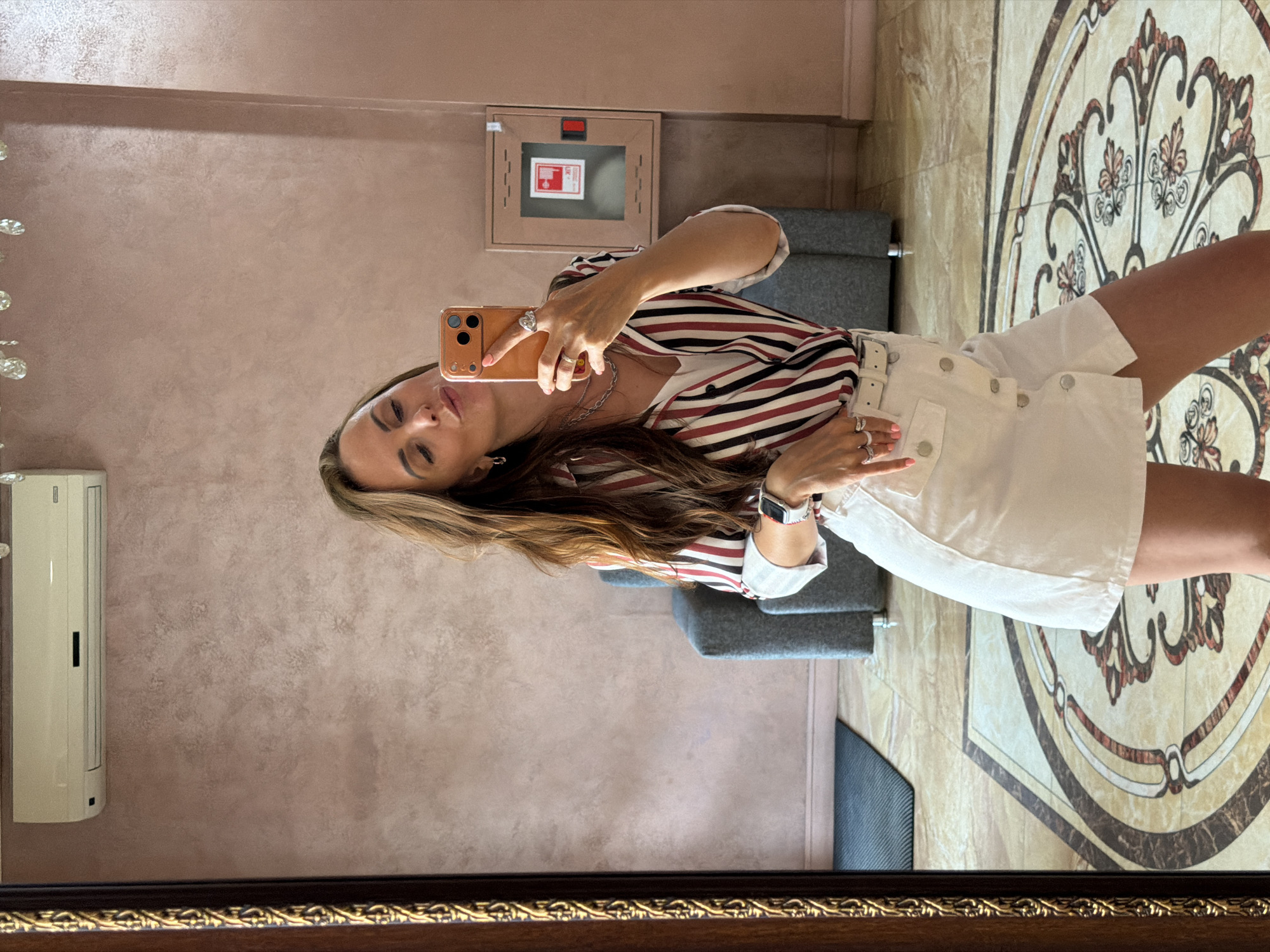 A woman is taking a mirror selfie in what appears to be a hotel lobby or a well-decorated room, possibly in Birobidzhan, Russia. She is wearing a white skirt with a belt and a vertically striped button-up shirt in red, white, and black. Her hair is long and brown, and she is wearing jewelry, including rings and a necklace. The background features a textured pink wall, an air conditioning unit, a fire extinguisher cabinet, a mirrored wall, a plush chair, and an ornate tiled floor. She is holding an orange smartphone to take the picture and has her lips puckered.