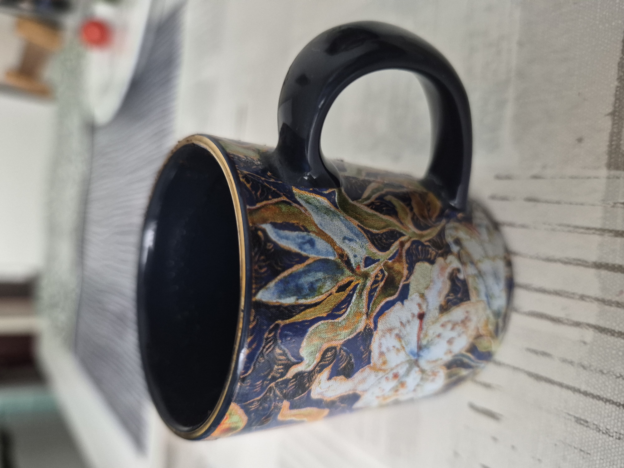 A close-up, eye-level shot shows a dark blue ceramic mug with a floral design. The mug has a gold rim and a dark blue handle. The floral pattern is intricate, featuring white and light blue flowers with accents of green and orange foliage. The mug is placed on a light-colored, textured surface with faint vertical lines. In the blurred background, a dark blue and white striped placemat and a white plate with a red object are visible. The scene is lit from above.
