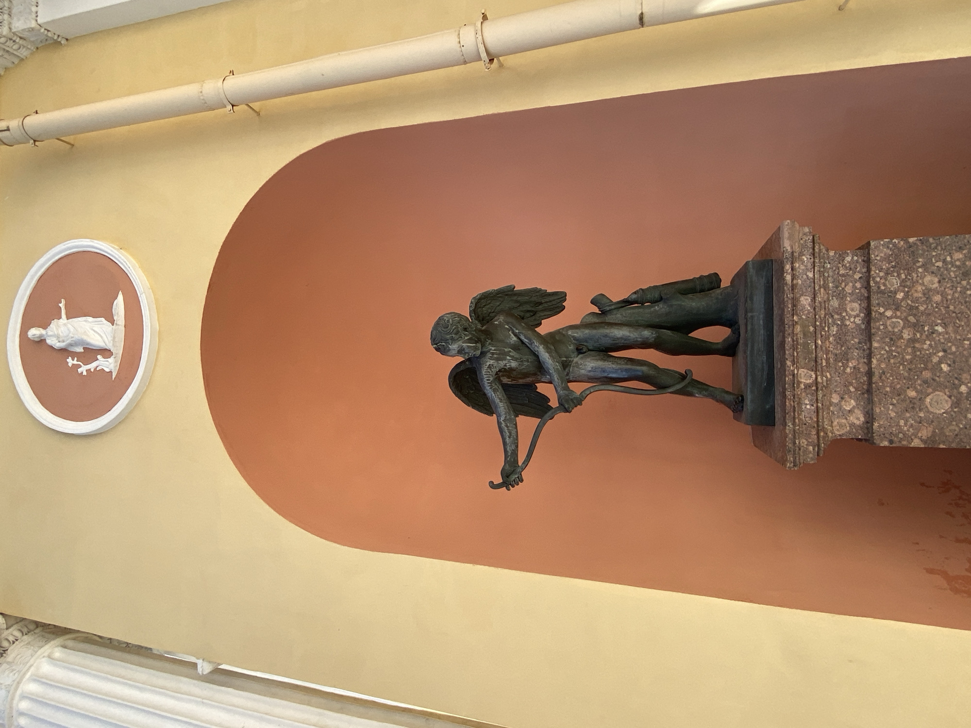 The image captures a statue of Cupid with a bow and arrow, standing on a granite pedestal in an alcove. The alcove is set within a building with a yellow facade. Above the alcove, a circular medallion with a bas-relief of a standing female figure is visible. A white fluted column stands to the left of the alcove, and a light-colored pipe runs vertically along the right side of the building. This scene is located in Pushkin, Russia, likely within the grounds of the Catherine Palace. The statue is made of dark metal, possibly bronze, and depicts Cupid in a dynamic pose, drawing his bow. The bas-relief above appears to be classical in style. The sunlight casts shadows, highlighting the architectural features and the sculpture.