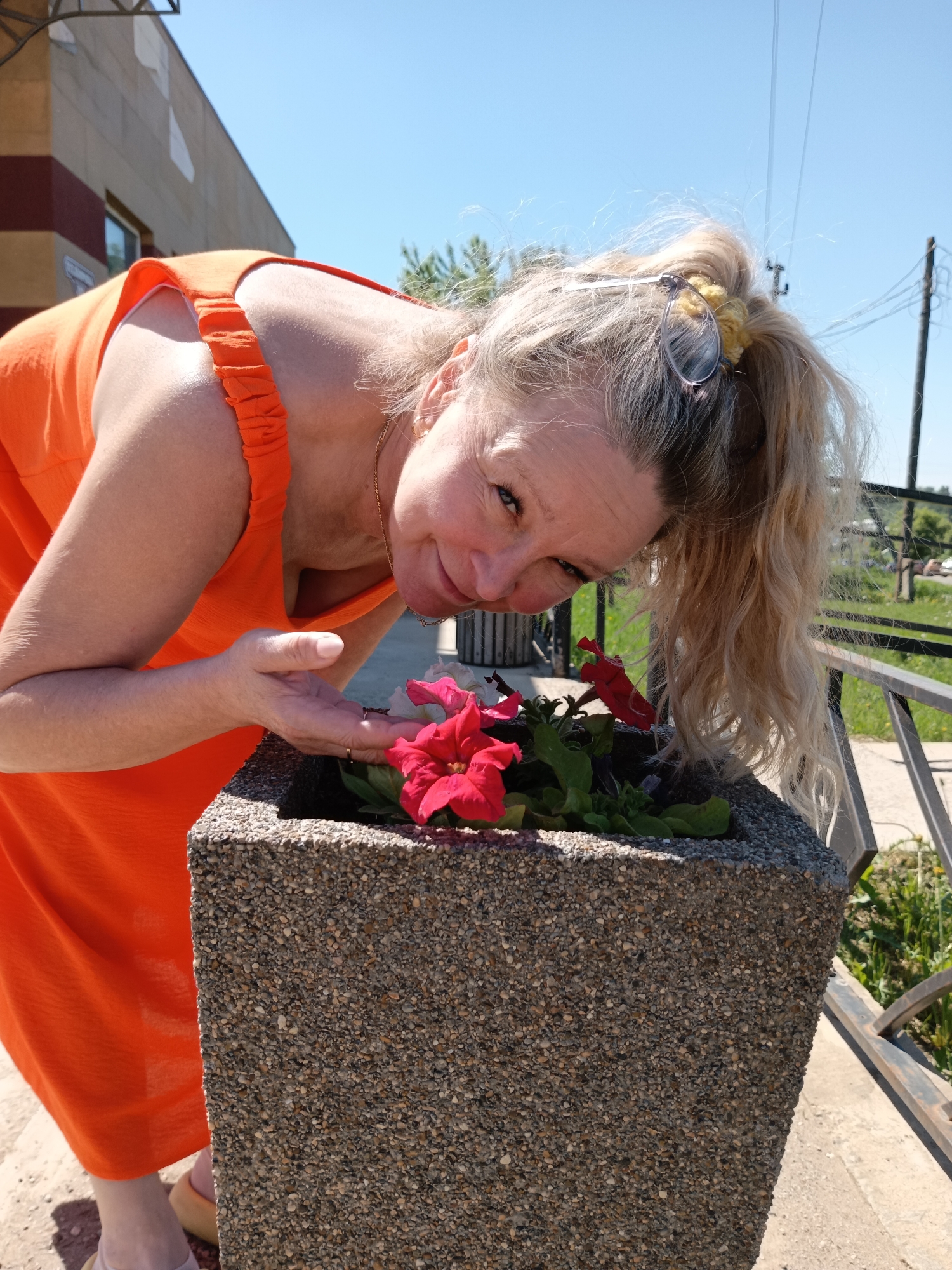The image shows a middle-aged woman in an orange dress leaning over a concrete planter box filled with red flowers. The woman has blonde hair pulled back with a yellow scrunchie, and she is looking at the camera with a slight smile. Her left hand is outstretched toward the flowers. The planter box is textured with small pebbles. In the background, there is a building with brown and red stripes, a metal railing, some greenery, and a clear blue sky. Based on the geolocation of Фоки, Russia, the scene is likely an outdoor space within the city.