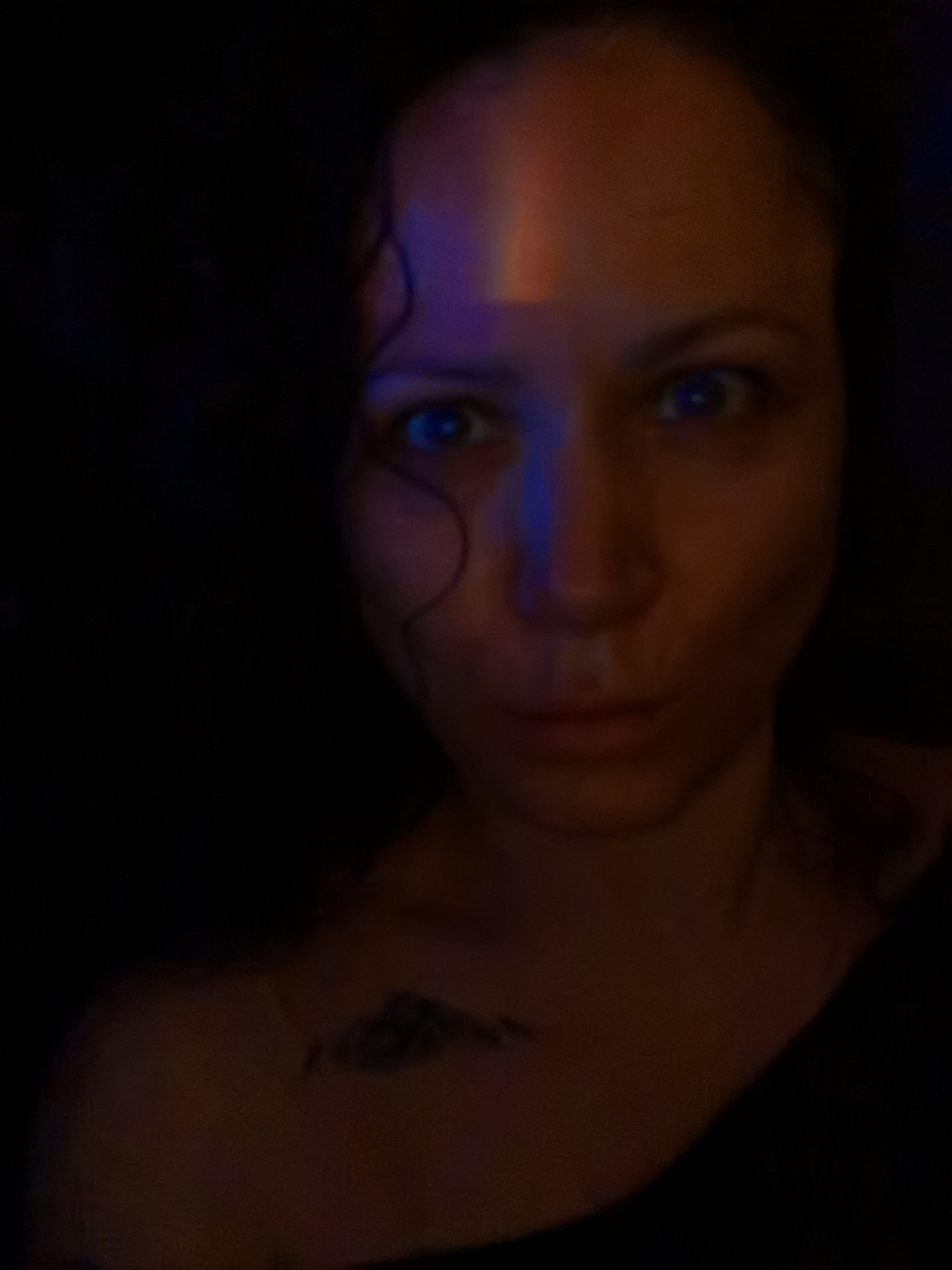 A close-up, blurred image shows a light-skinned individual with blue eyes looking directly at the camera. A rainbow light projection is visible on their forehead and nose. The person has dark hair and a tattoo is visible on their upper chest. The background is dark and indistinct.