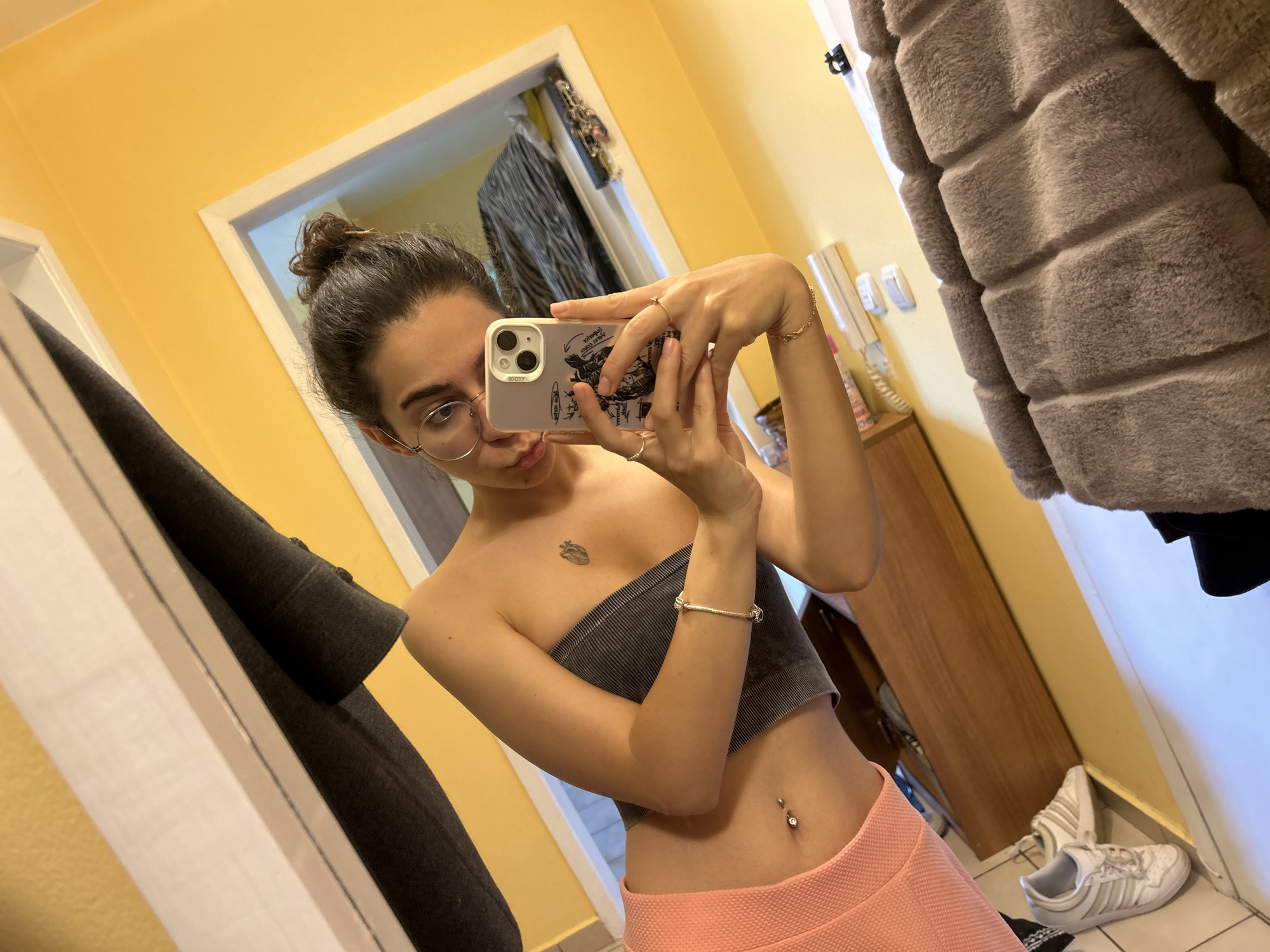 A young woman with dark hair in a bun and glasses is taking a selfie in a room. She is wearing a grey strapless top and pink shorts, with a belly button piercing. A tattoo of a heart is visible on her chest. She is holding up a smartphone to take the picture. In the background, there is a wooden dresser, a fur coat hanging on a hook, and a pair of white sneakers on the floor. The walls are painted yellow. The location is not identifiable as Sofia, Bulgaria.