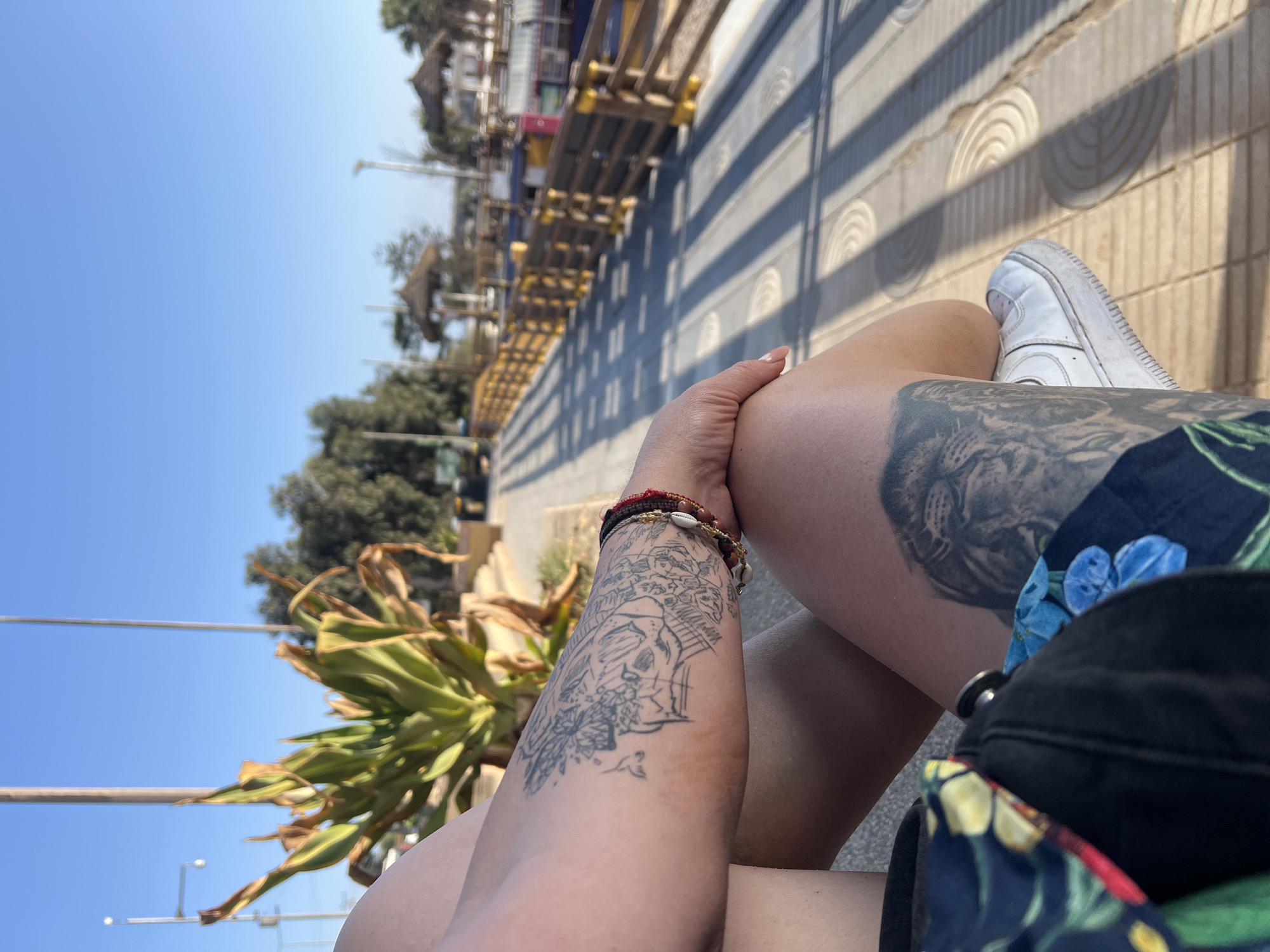 A medium-angle, eye-level outdoor shot shows a woman sitting in an urban setting, likely in Arica, Chile. The location is recognizable through the specific architectural elements and landscaping visible in the background. The woman's crossed legs dominate the foreground. Tattoos are visible on both her arms and upper thigh. The left arm features a detailed black and white skull design. Her right upper thigh has a tattoo of a lion's face. A casual black tote bag with a floral design sits on her lap. A single white sneaker rests on the concrete surface. A line of construction and buildings are along the top left. A line of trees is on the lower left. In the background, a low stone wall with a wave pattern casts shadows across the concrete. The weather appears sunny, as indicated by the bright, clear blue sky and sharp shadows.