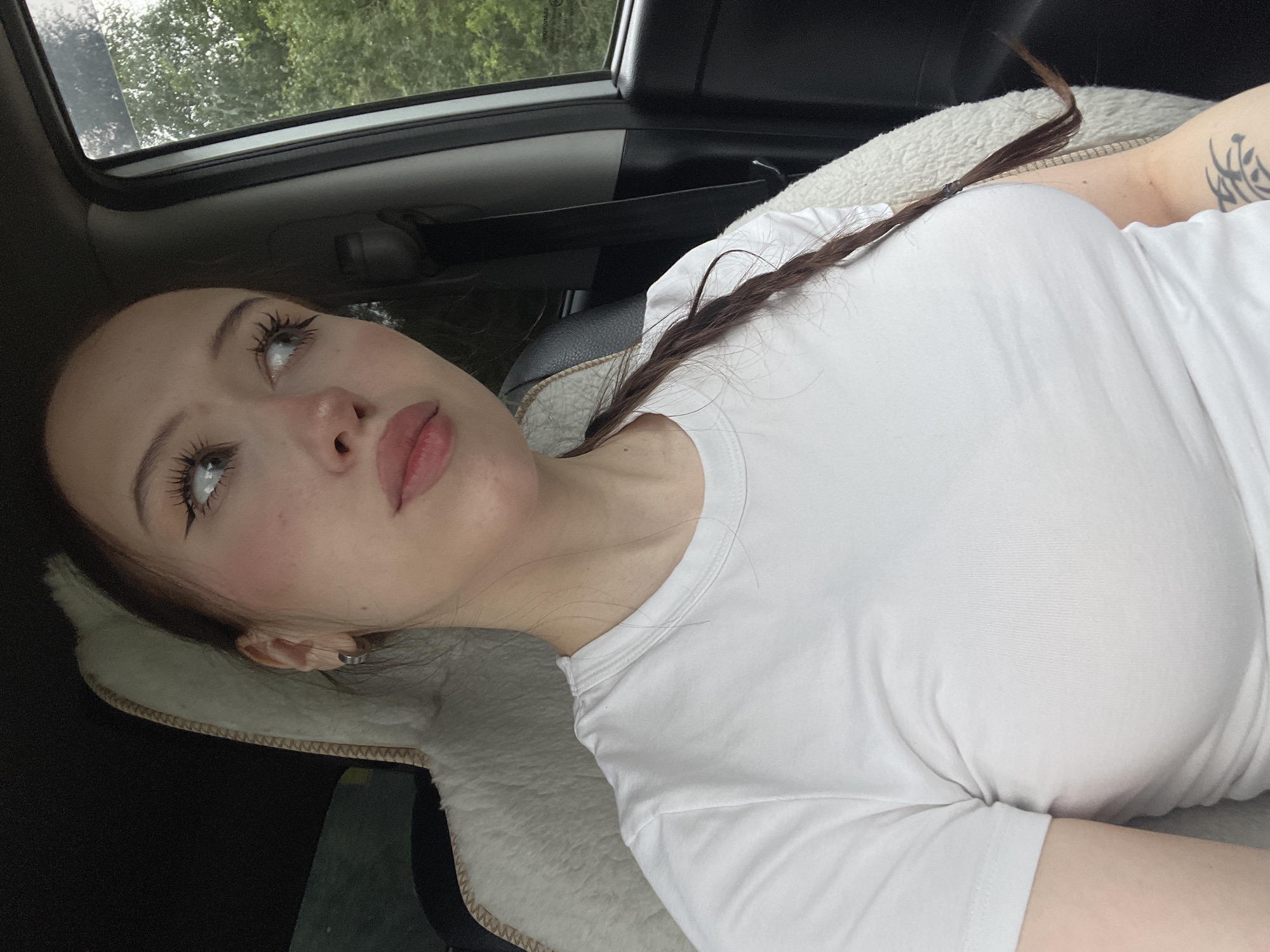 A young woman with long brown hair in a braid and heavy eye makeup is sitting in the passenger seat of a car. She is wearing a white t-shirt and looking out the window. The interior of the car is visible, including a fluffy seat cover and a black car seat. Outside the window, trees and greenery can be seen, suggesting a rural or natural setting. The lighting indicates it is daytime. There are no visible indicators suggesting this specific location is Gavrilovka, Russia.