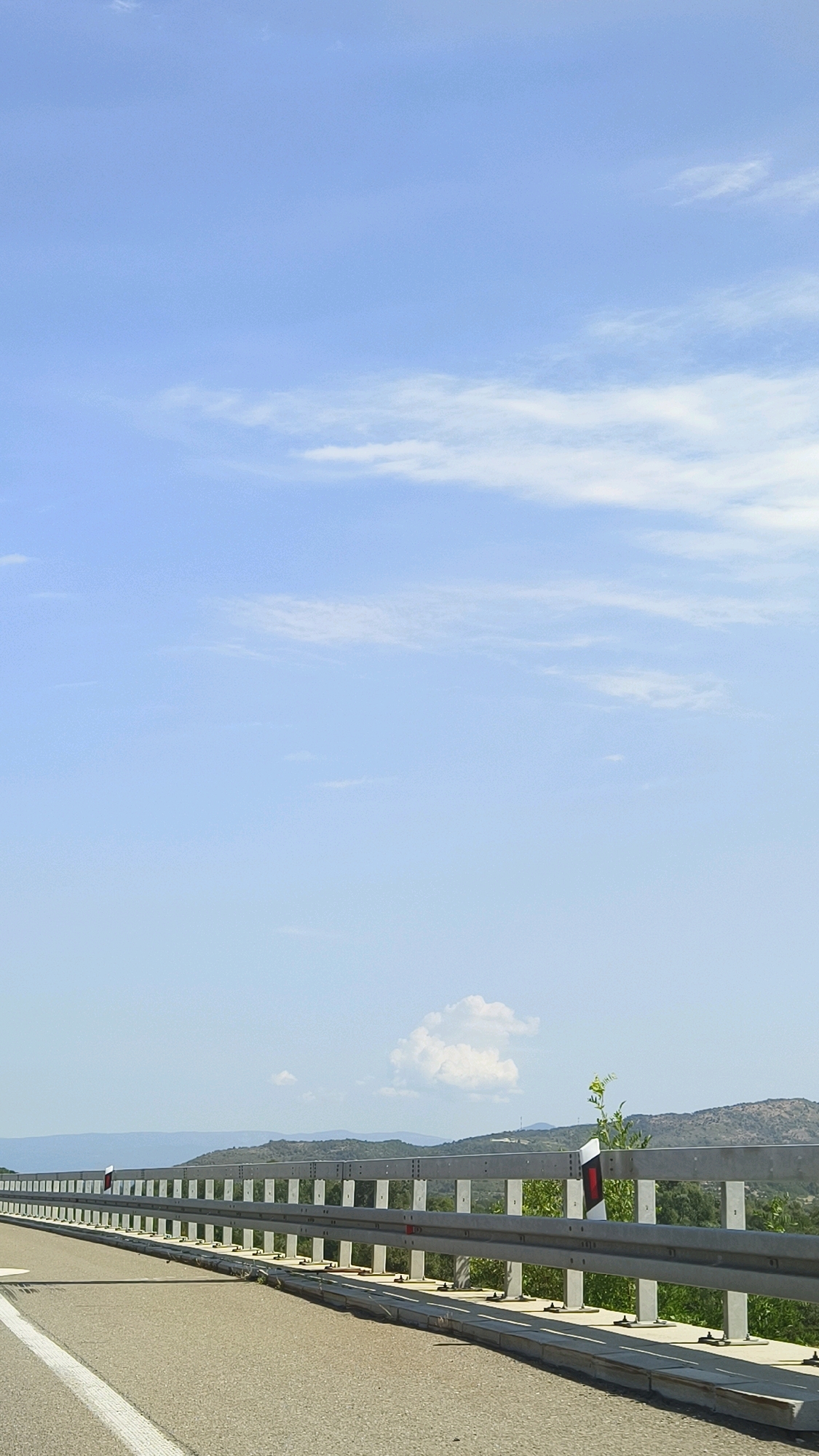 The image shows a highway guardrail running along the side of a road. The road is paved and has white lane markings. The guardrail is made of metal and has reflective markers. In the background, there are rolling hills covered in vegetation. The sky is clear and blue with a few scattered clouds. This appears to be a scene on the E761 highway near Vranje, Serbia, a major route that connects various cities across the country, including Vranje to Niš. The image captures a typical stretch of rural highway, likely intended for travel or transport.