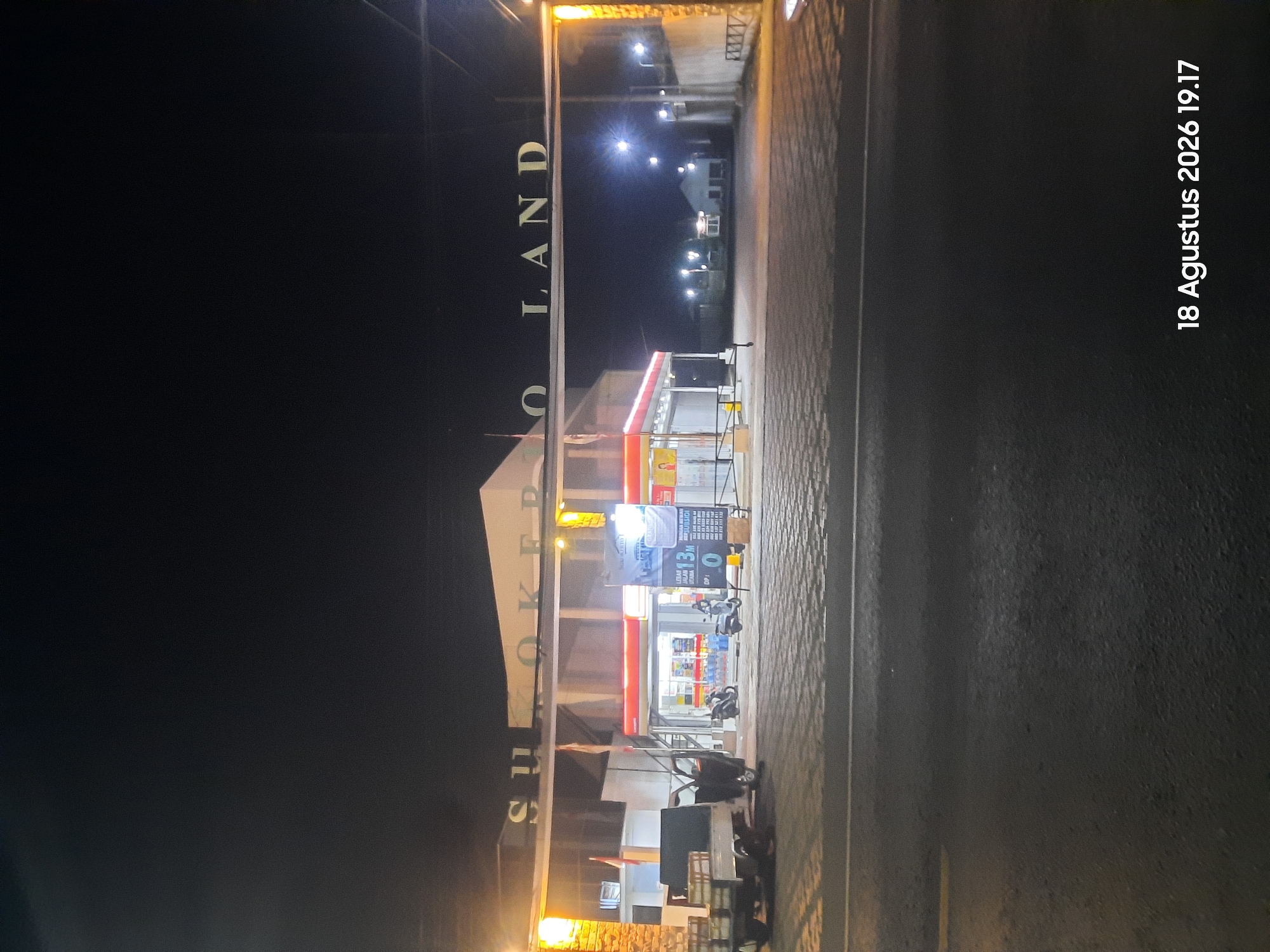 The image shows an entrance gate with the text "SUKOKERTO LAND" at night in Sukokerto, Indonesia. A convenience store with a red and white awning is visible behind the gate. Several motorcycles are parked in front of the store. A small truck is parked to the left of the entrance. The ground in front of the store is paved with interlocking bricks, and the road in the foreground is asphalt. Streetlights are visible in the background, illuminating some buildings and trees. A digital timestamp in the bottom right corner reads "18 Agustus 2026 19.17".